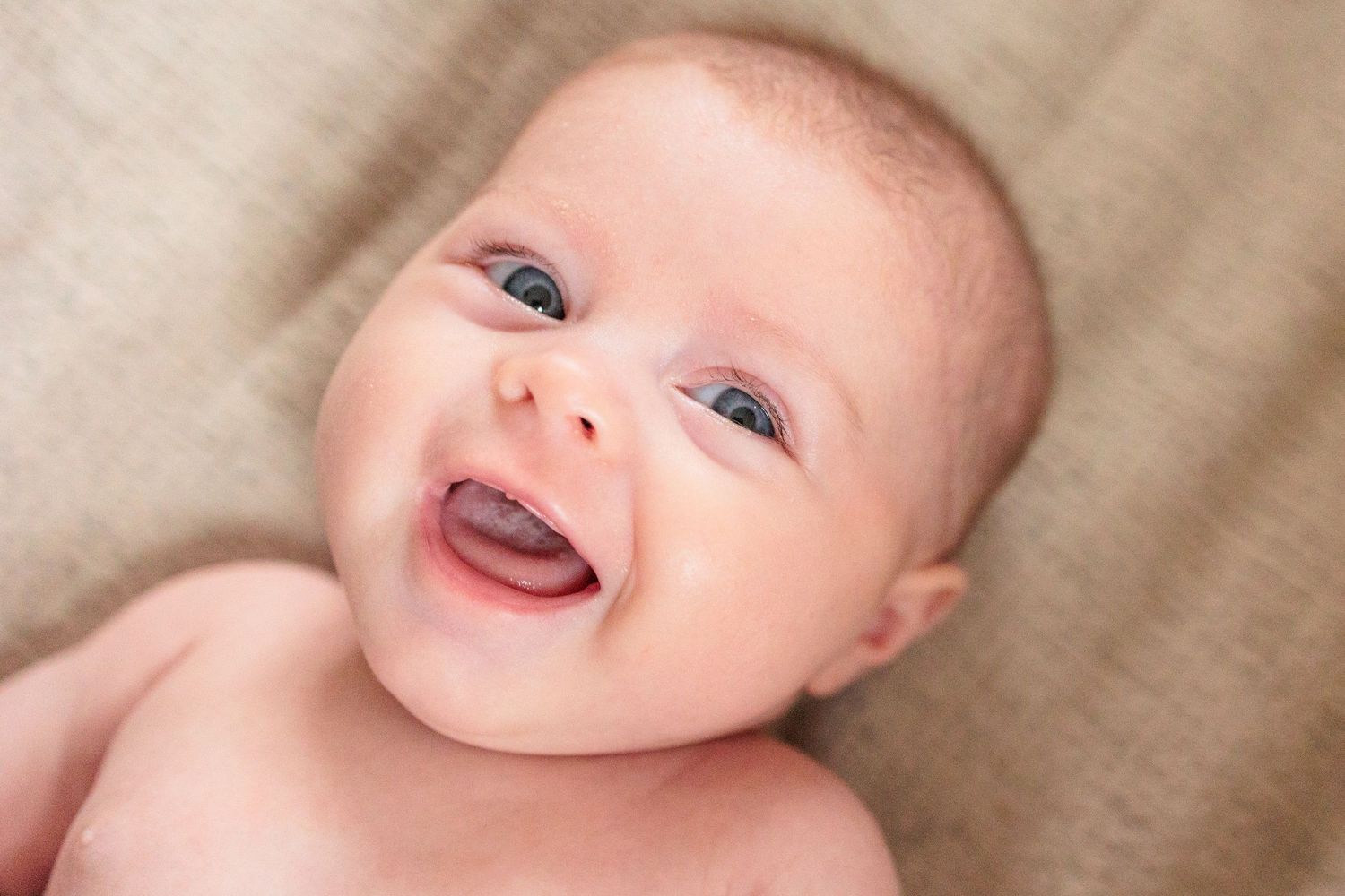 Close up view of a baby with bright blue eyes and rosy cheeks lying on beige fabric.