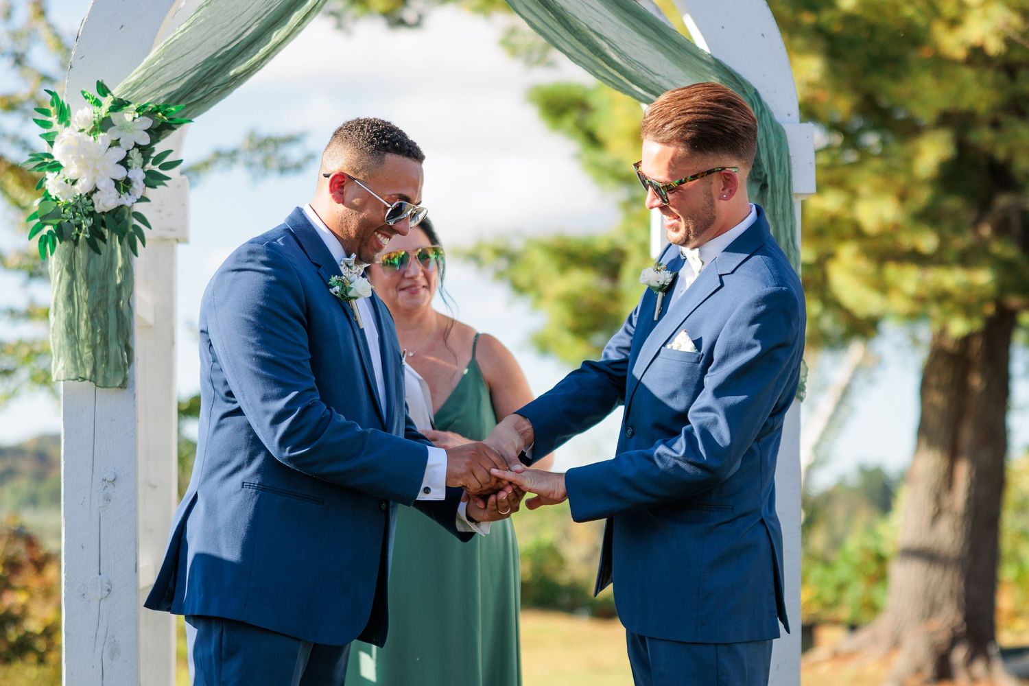 Wedding ceremony participants in blue suits gather under a decorated white arch outdoors.