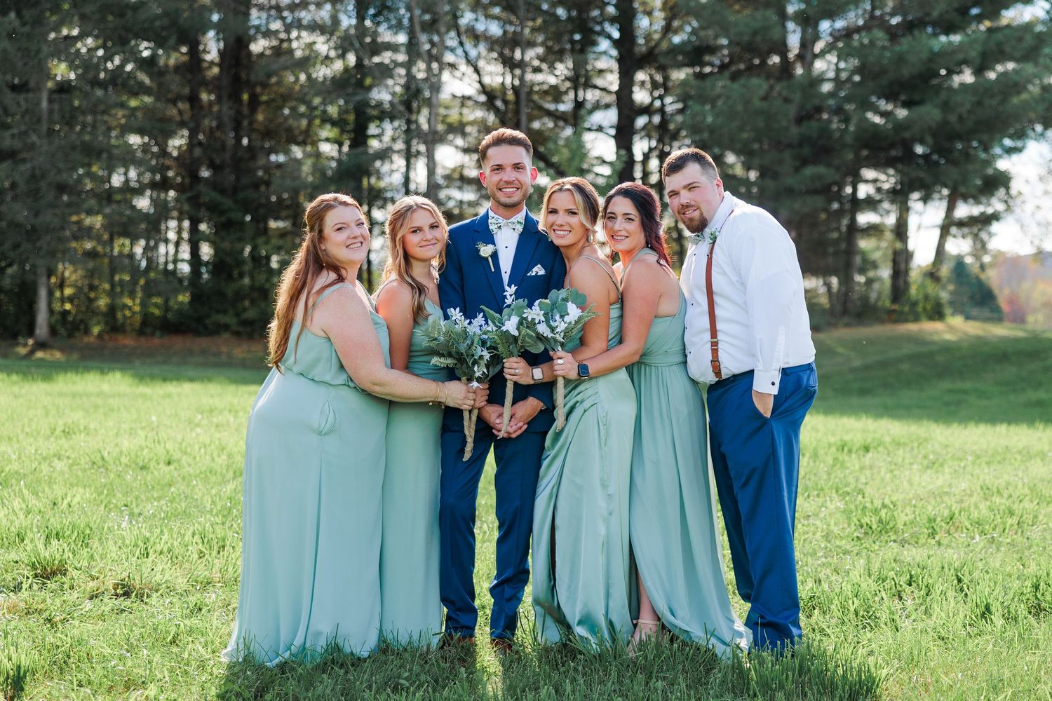 Wedding party in coordinating mint green dresses and navy suits pose playfully on grass with pine trees behind.