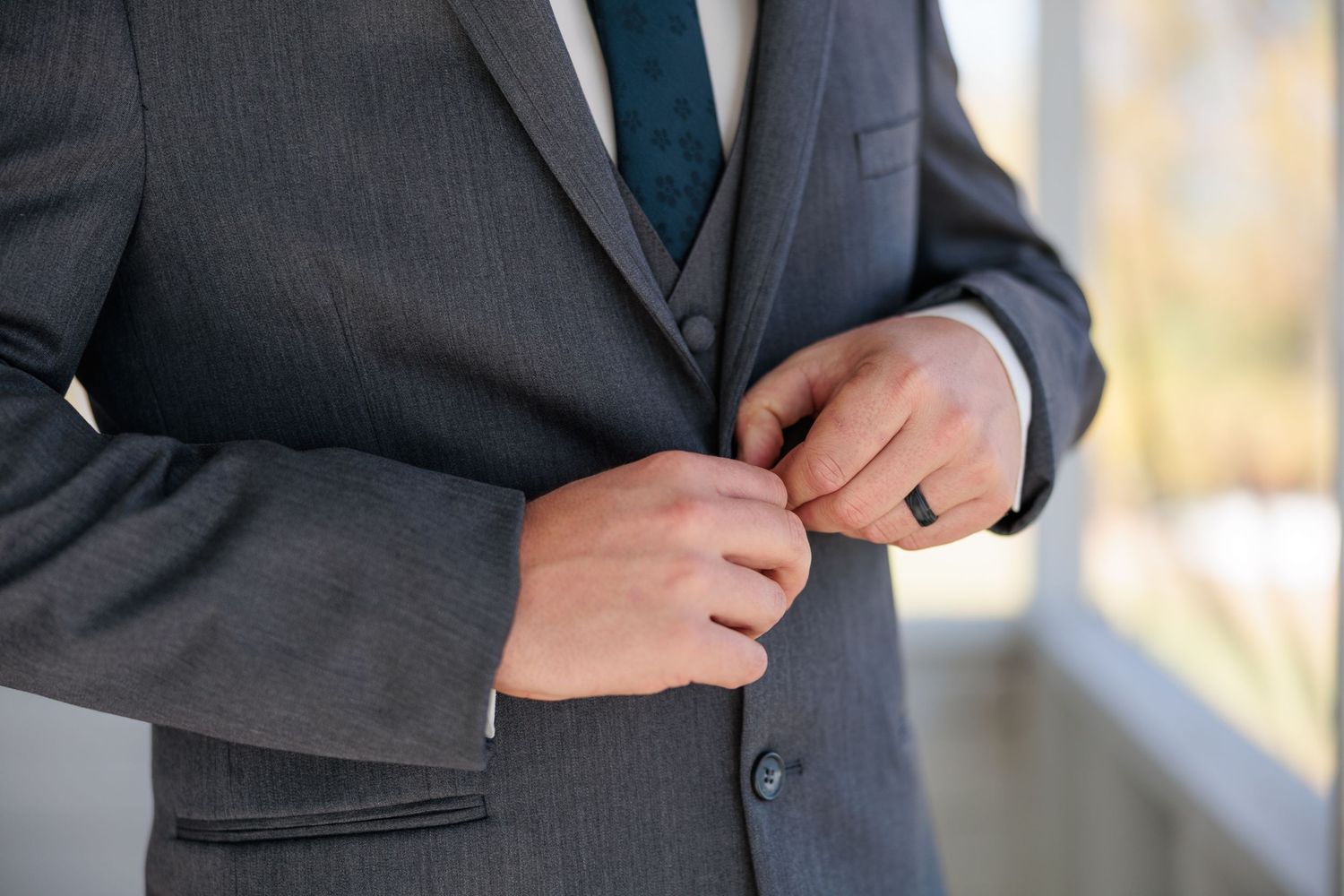 Close-up of groom in gray three-piece suit fastening jacket button, wearing black wedding band.