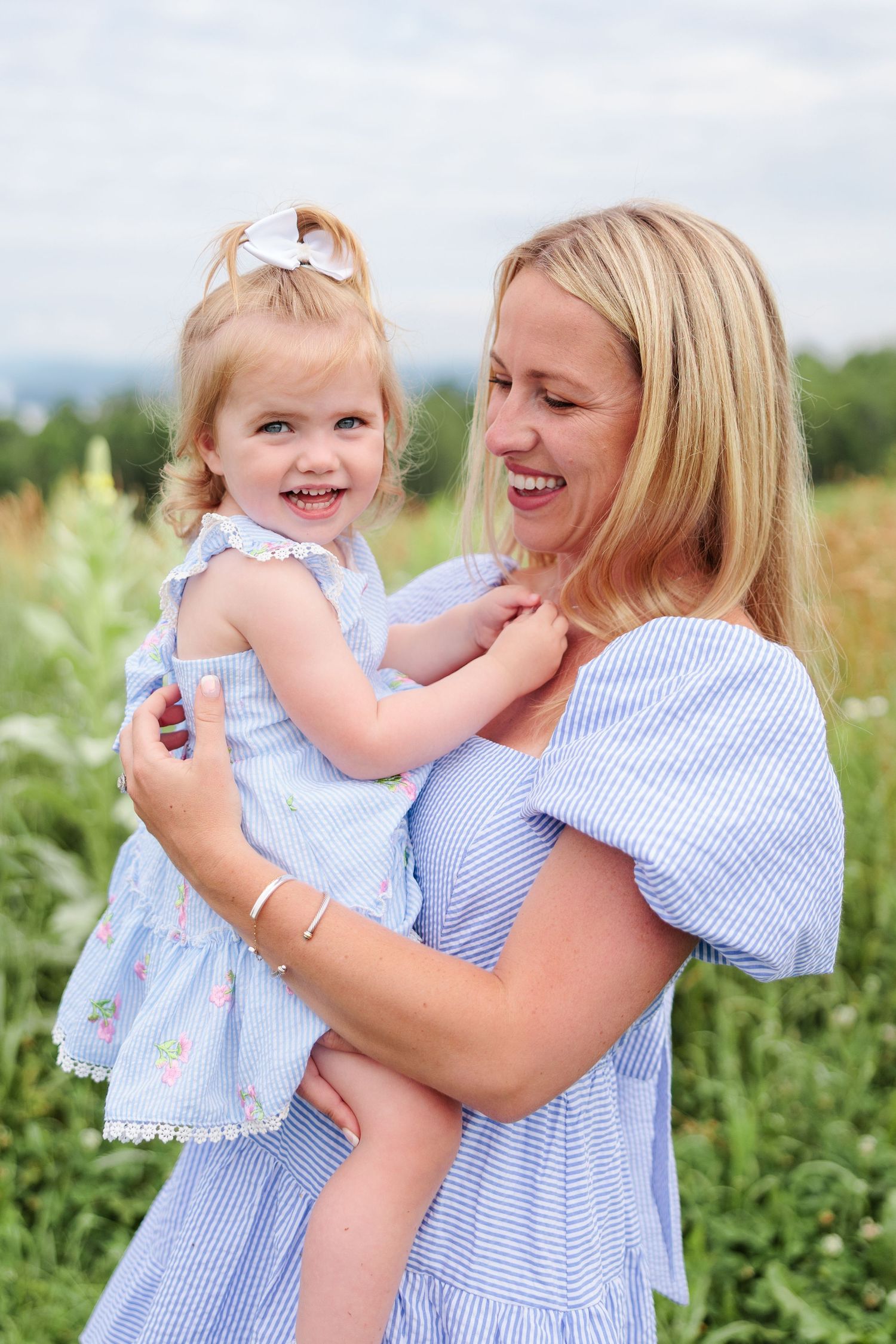 Laughing mother and daughter in matching blue dresses enjoying a sunny day in a flower field.