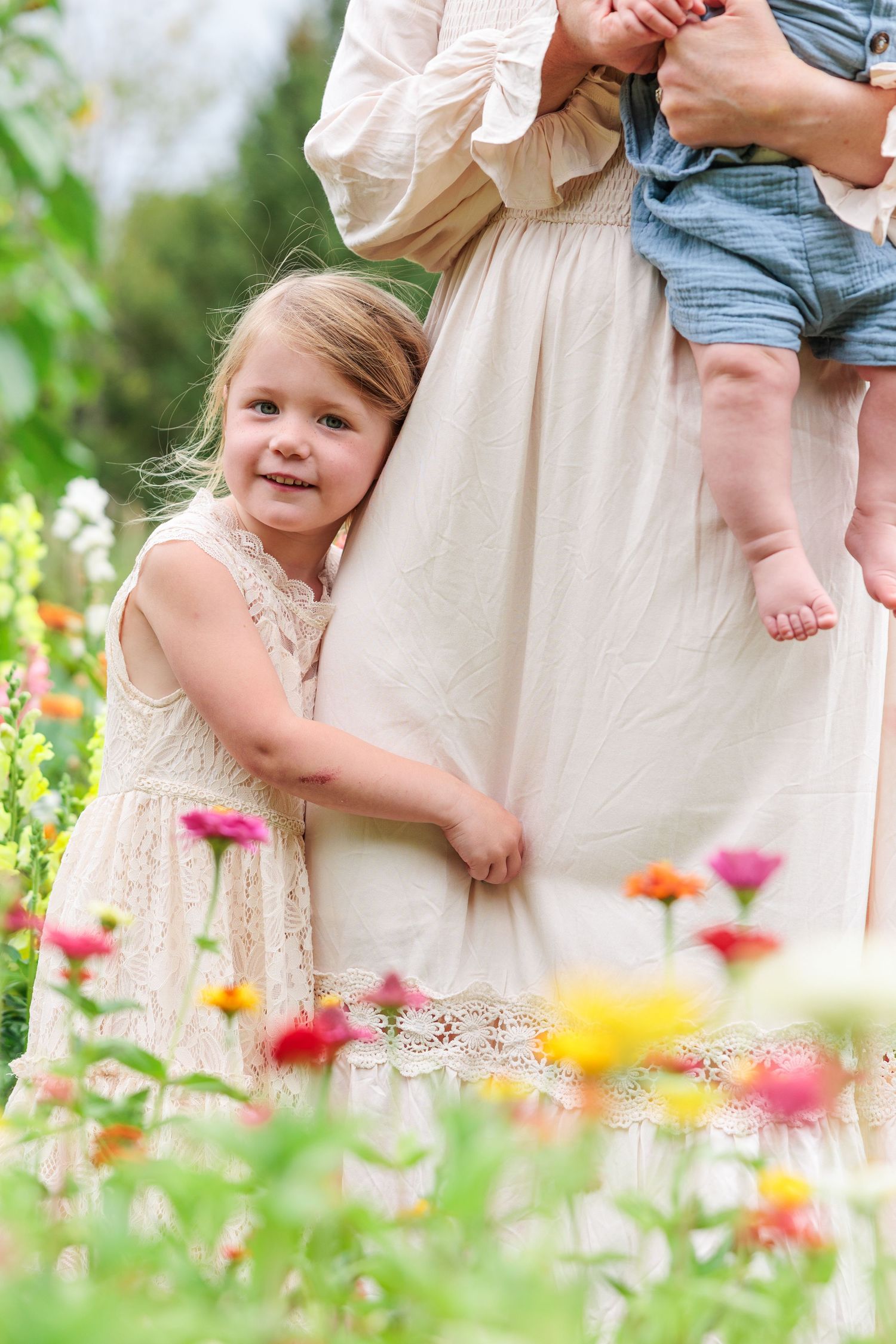 A young girl in a lace dress hugs her mother's skirt in a colorful flower garden.