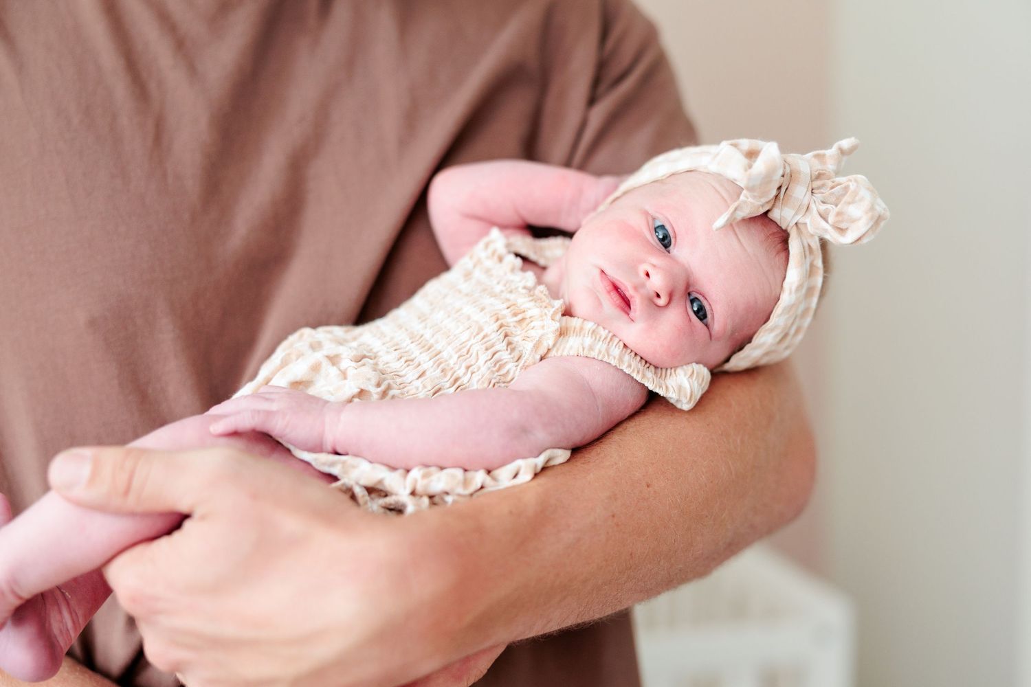 Adorable newborn baby girl in lace outfit and bow headband cradled in fathers arms.