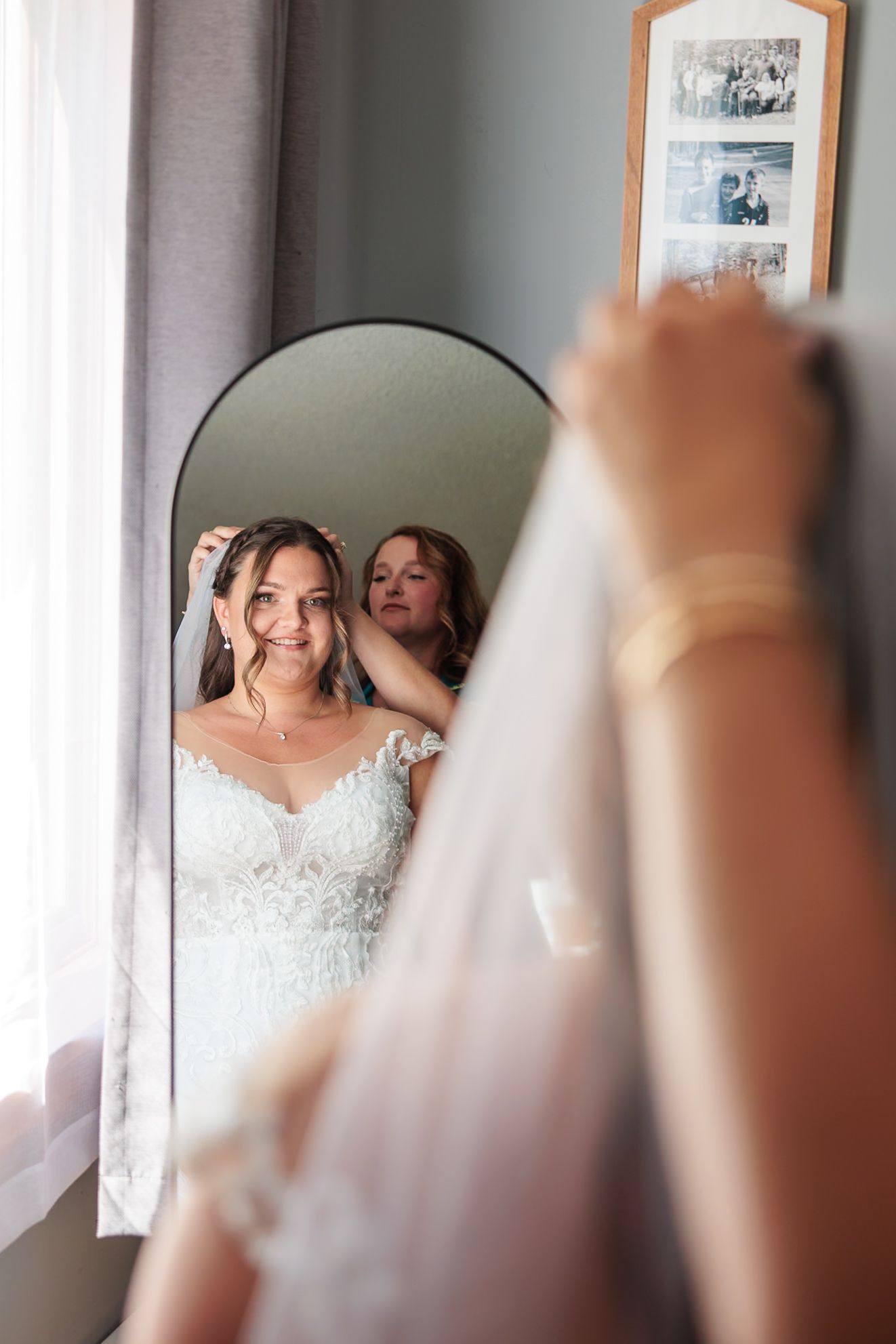 Bridesmaid helps bride with veil while smiling bride looks in mirror.