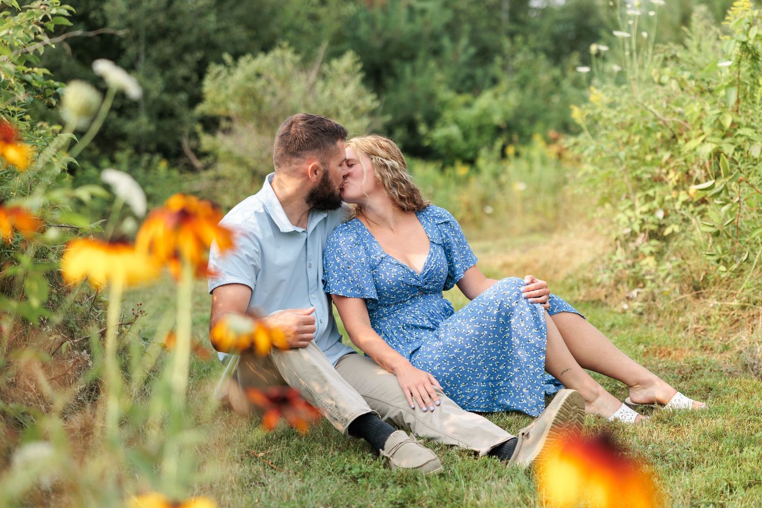 Romantic couple sharing a kiss while sitting in green grass surrounded by vibrant summer wildflowers.