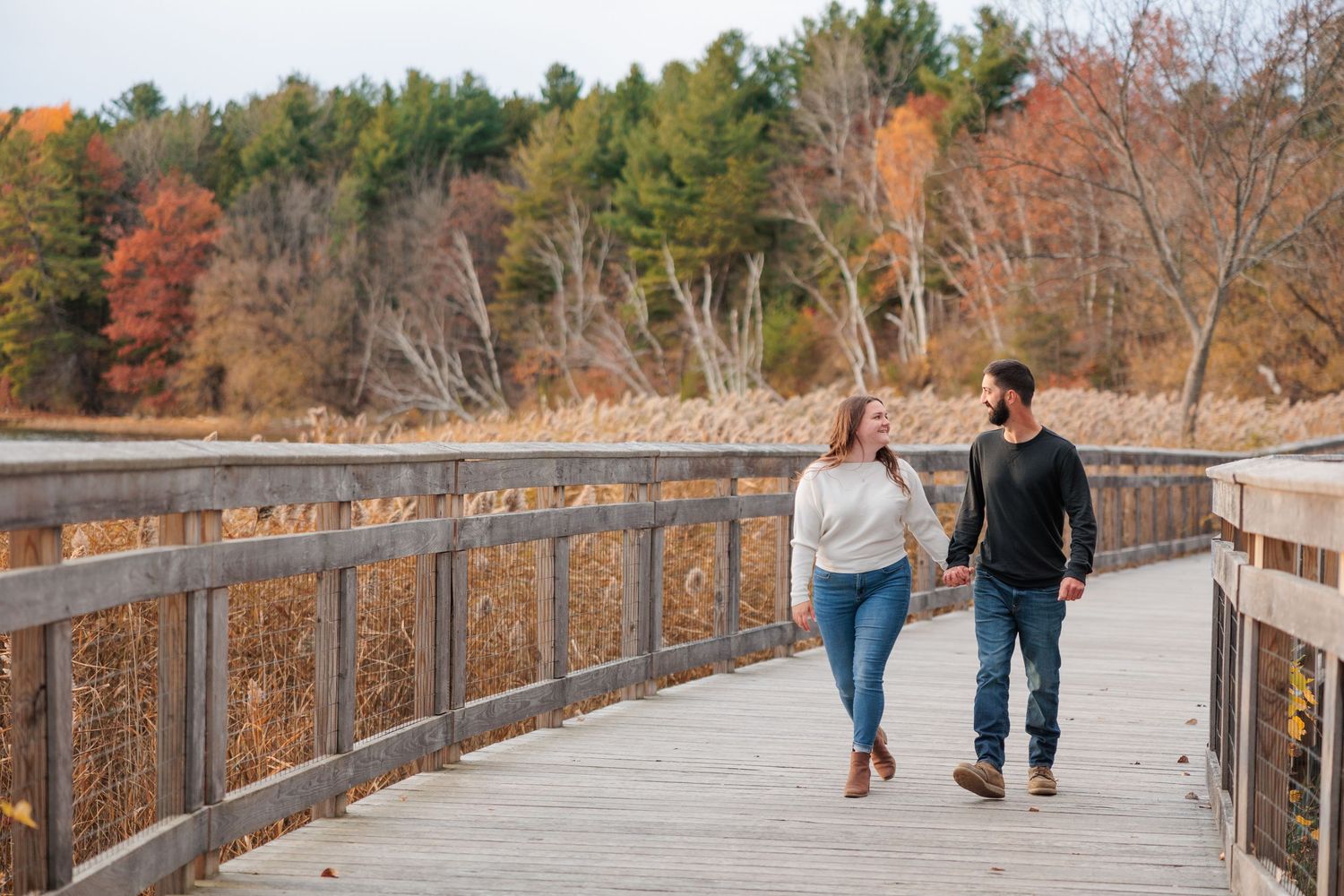 Couple holding hands walking on wooden boardwalk bridge surrounded by colorful fall foliage and wetlands.