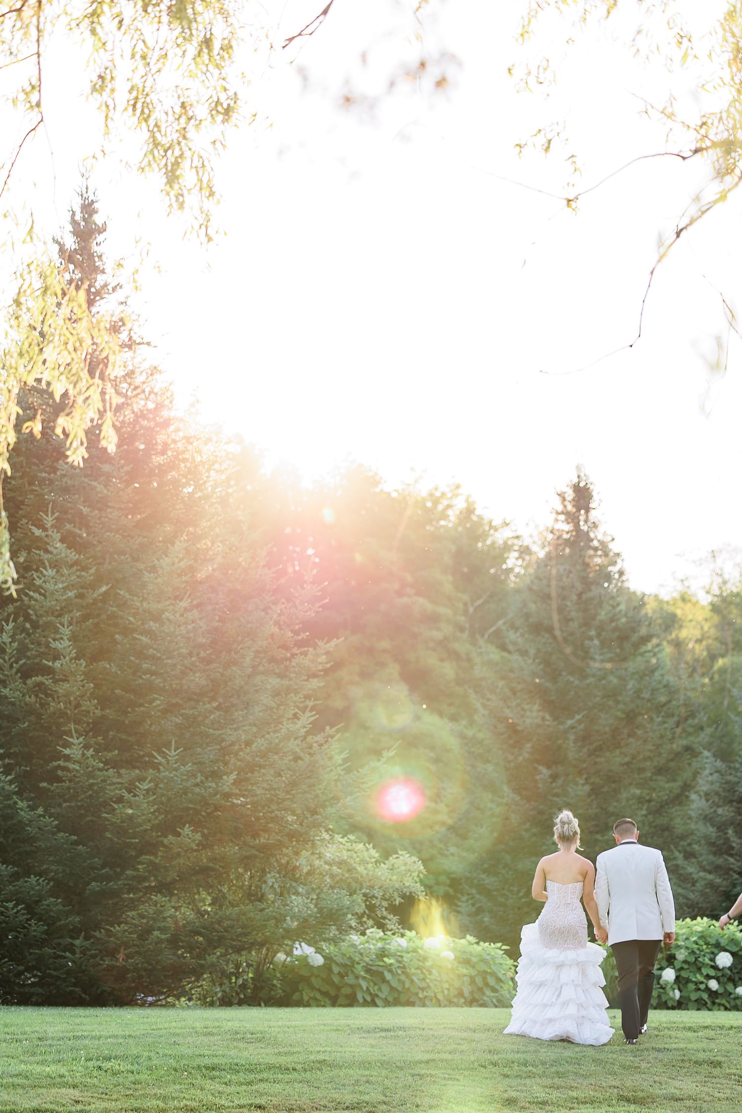 Bride and groom walking hand in hand toward stunning golden sunset through lush green garden landscape.