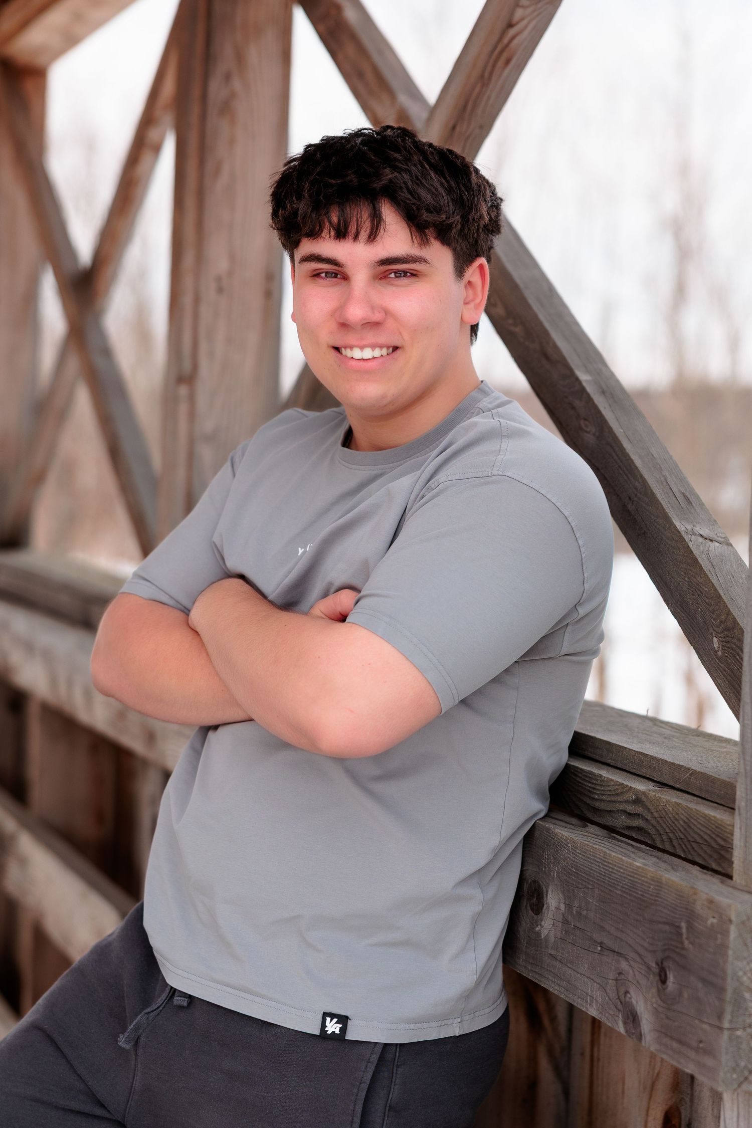 Smiling young man with dark hair, arms crossed, leaning on a wooden bridge railing wearing a gray t-shirt.
