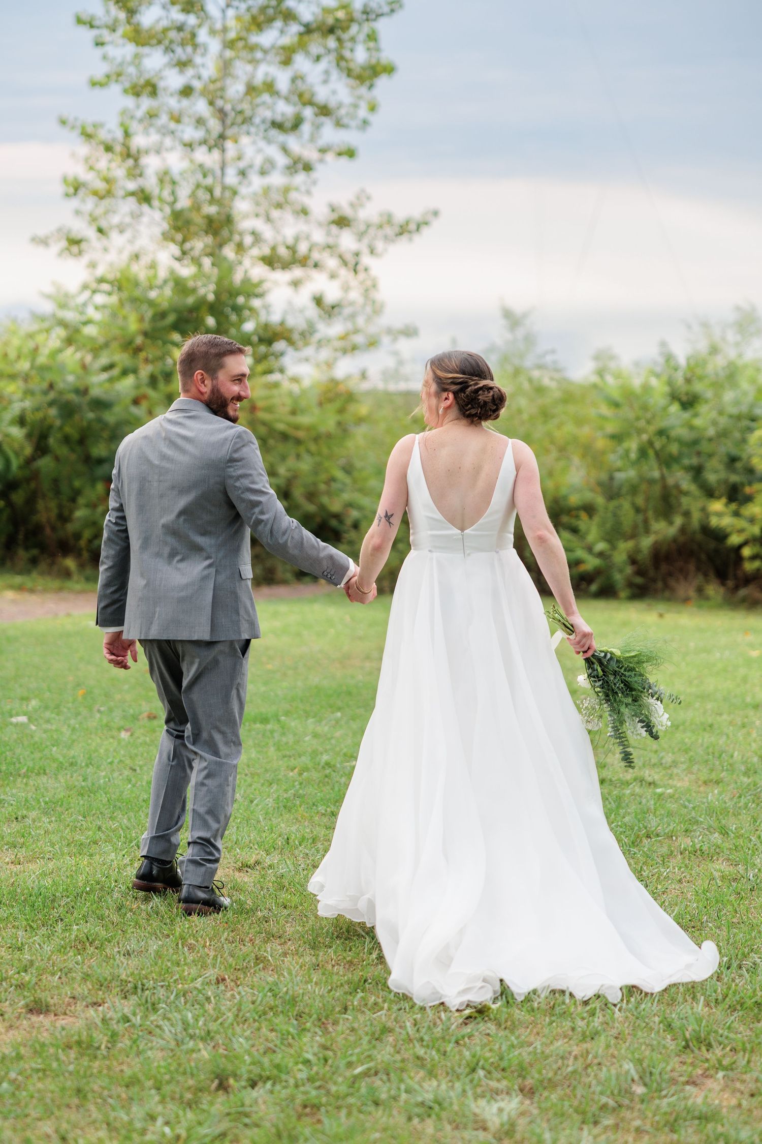 Bride and groom holding hands walking through green garden, groom smiling back at bride in white gown.