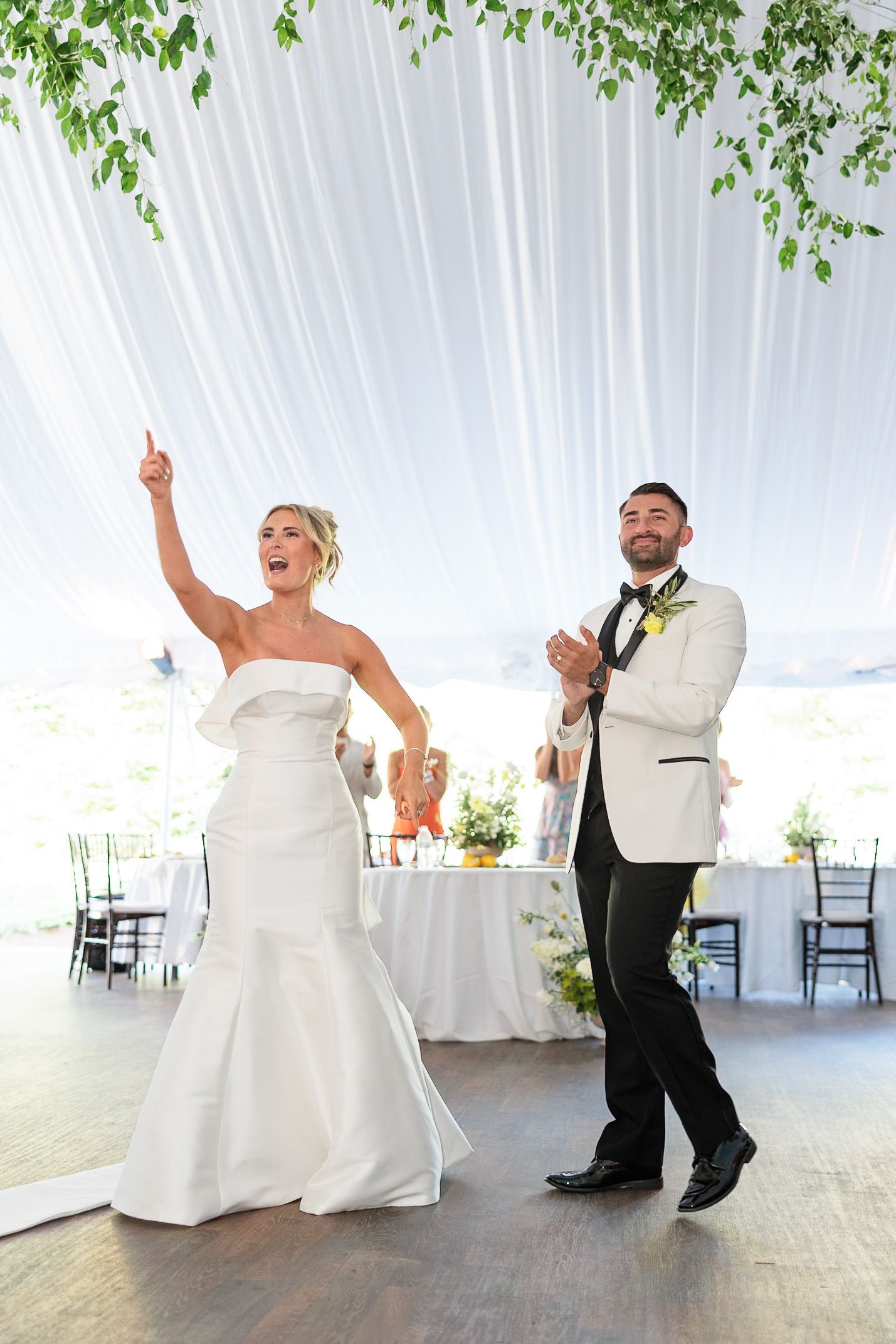Bride and groom make grand entrance onto dance floor under white draped ceiling with greenery.