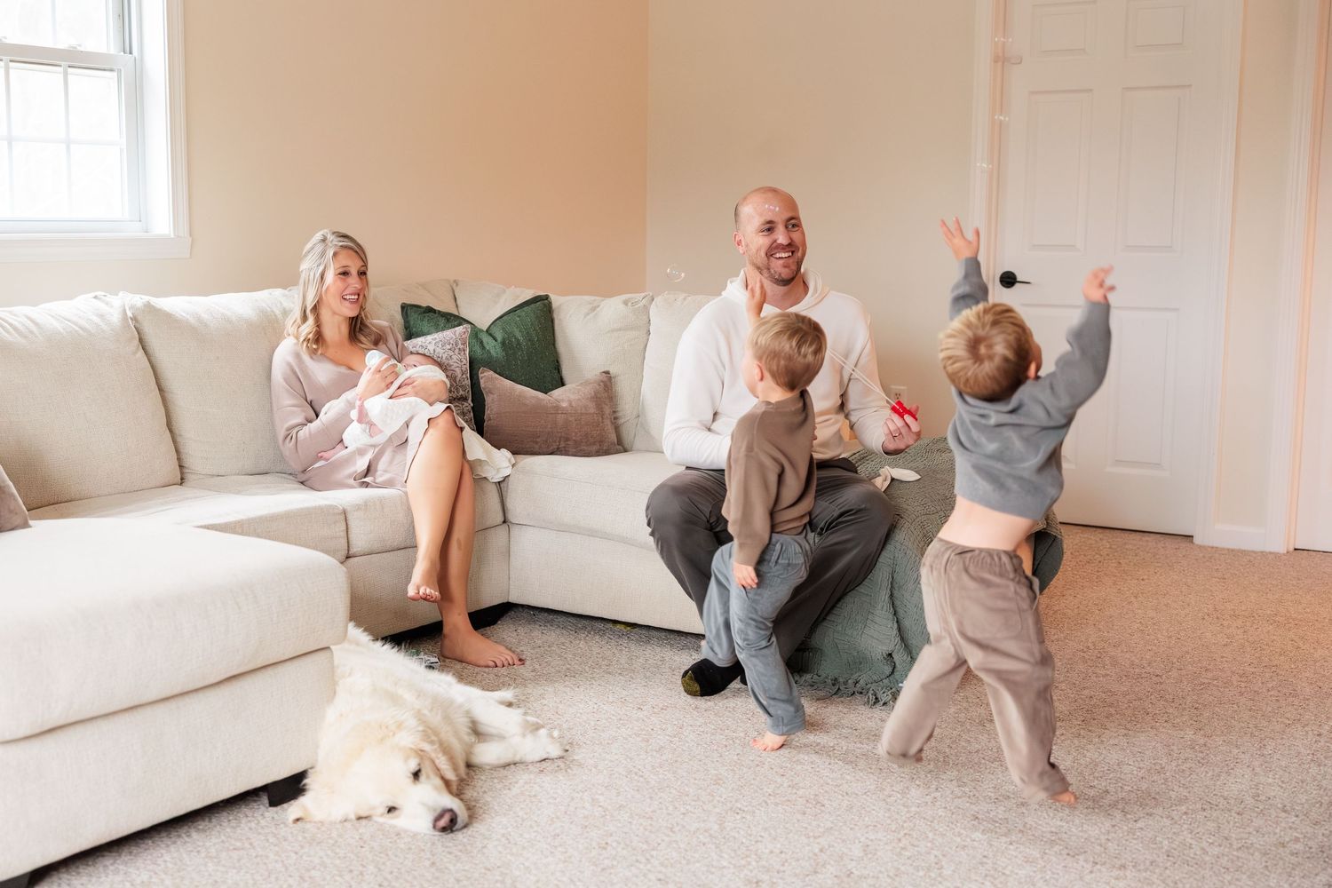 Happy family with newborn, two young boys, and golden retriever enjoying time together in bright living room.