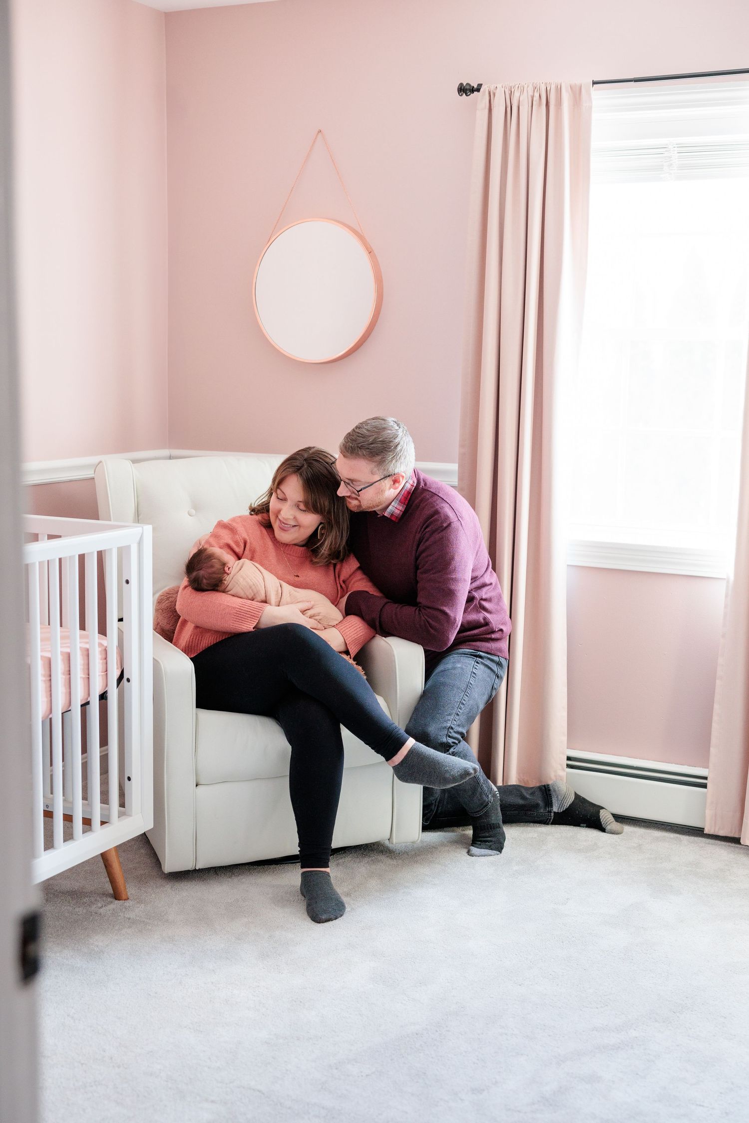 Parents sitting in nursery chair together holding newborn in pink decorated baby room.