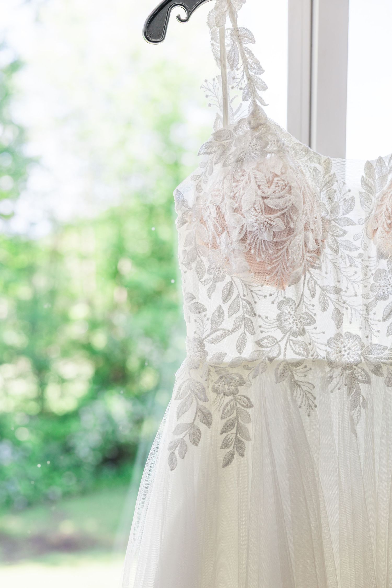 Close-up of an elegant lace and tulle wedding gown with floral embroidery hanging by a sunlit window.
