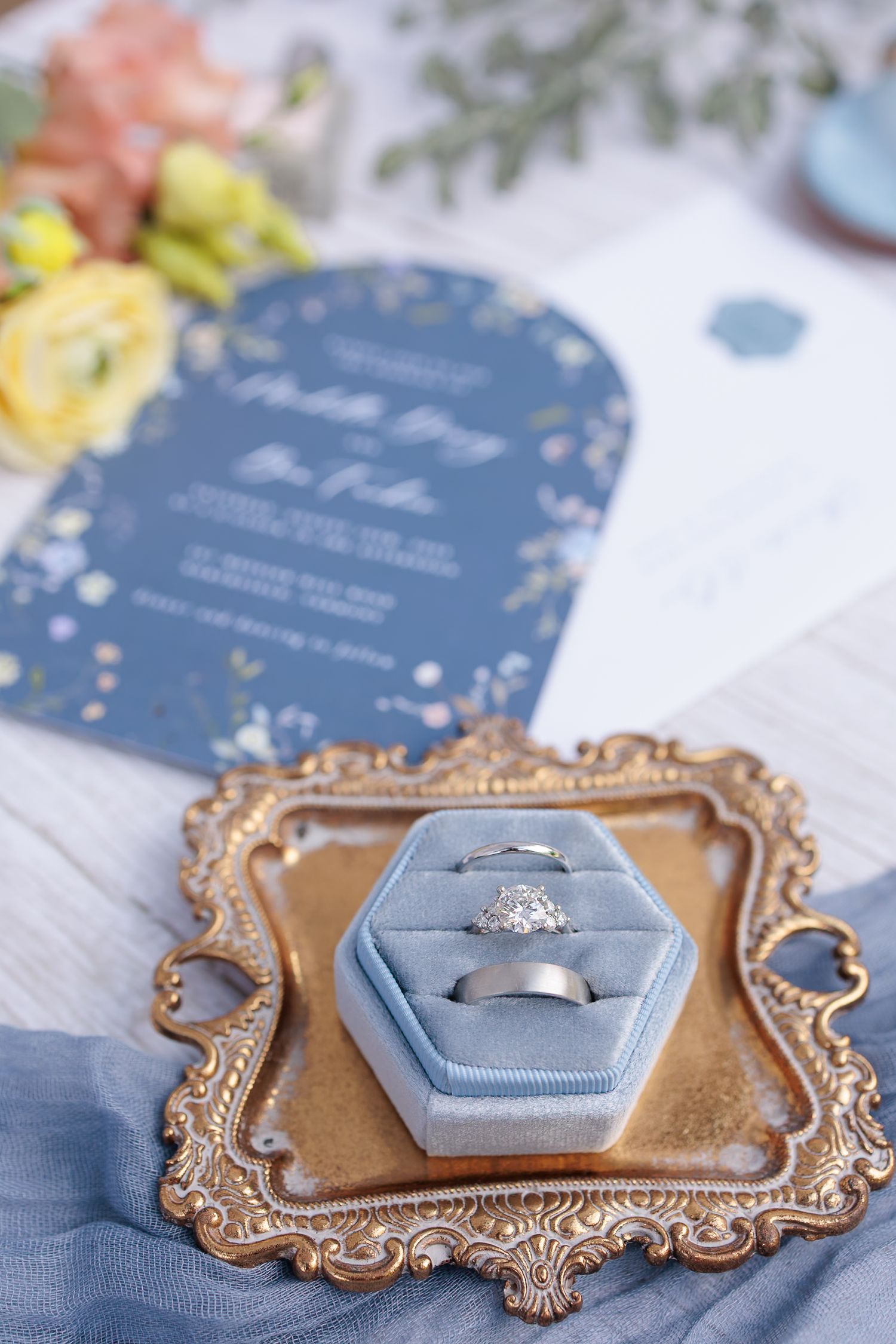 Wedding rings in a dusty blue velvet hexagon ring box on an ornate gold tray beside a floral invitation.