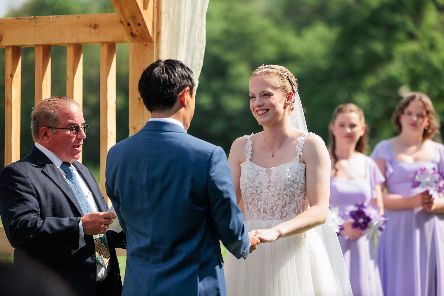 Bride laughing joyfully while exchanging vows with groom at outdoor wedding ceremony with bridesmaids.