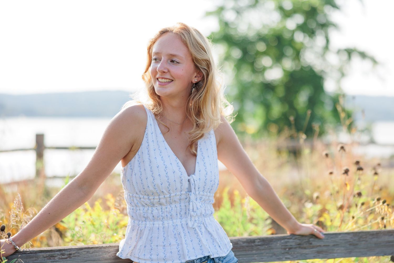 Young blonde woman smiling outdoors near a wooden fence with a scenic waterfront background on a sunny day.