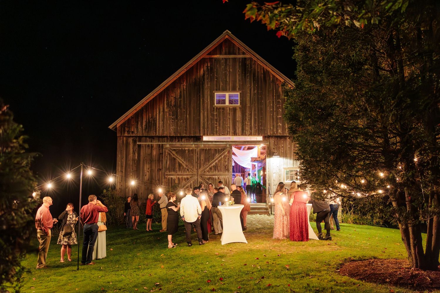 Wedding guests celebrate outside a wooden barn decorated with warm string lights in the evening.