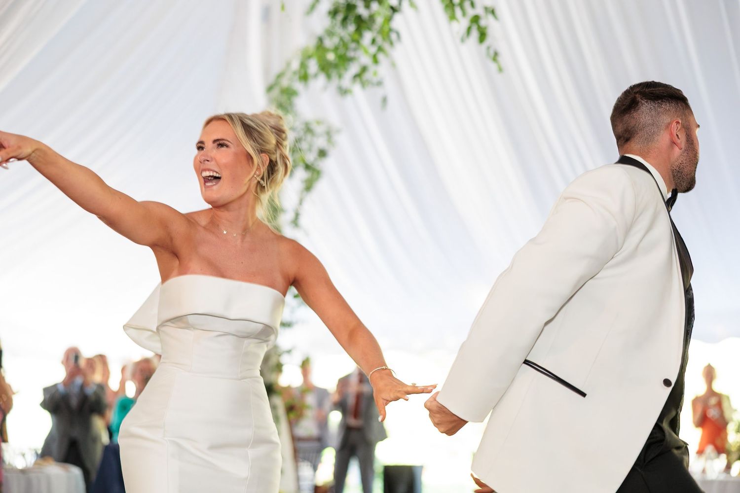 Bride and groom joyfully enter wedding reception under elegant white draped tent during first dance.
