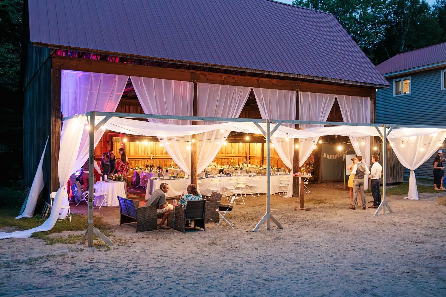 Elegant outdoor barn wedding reception with draped white curtains and warm string lights at dusk.