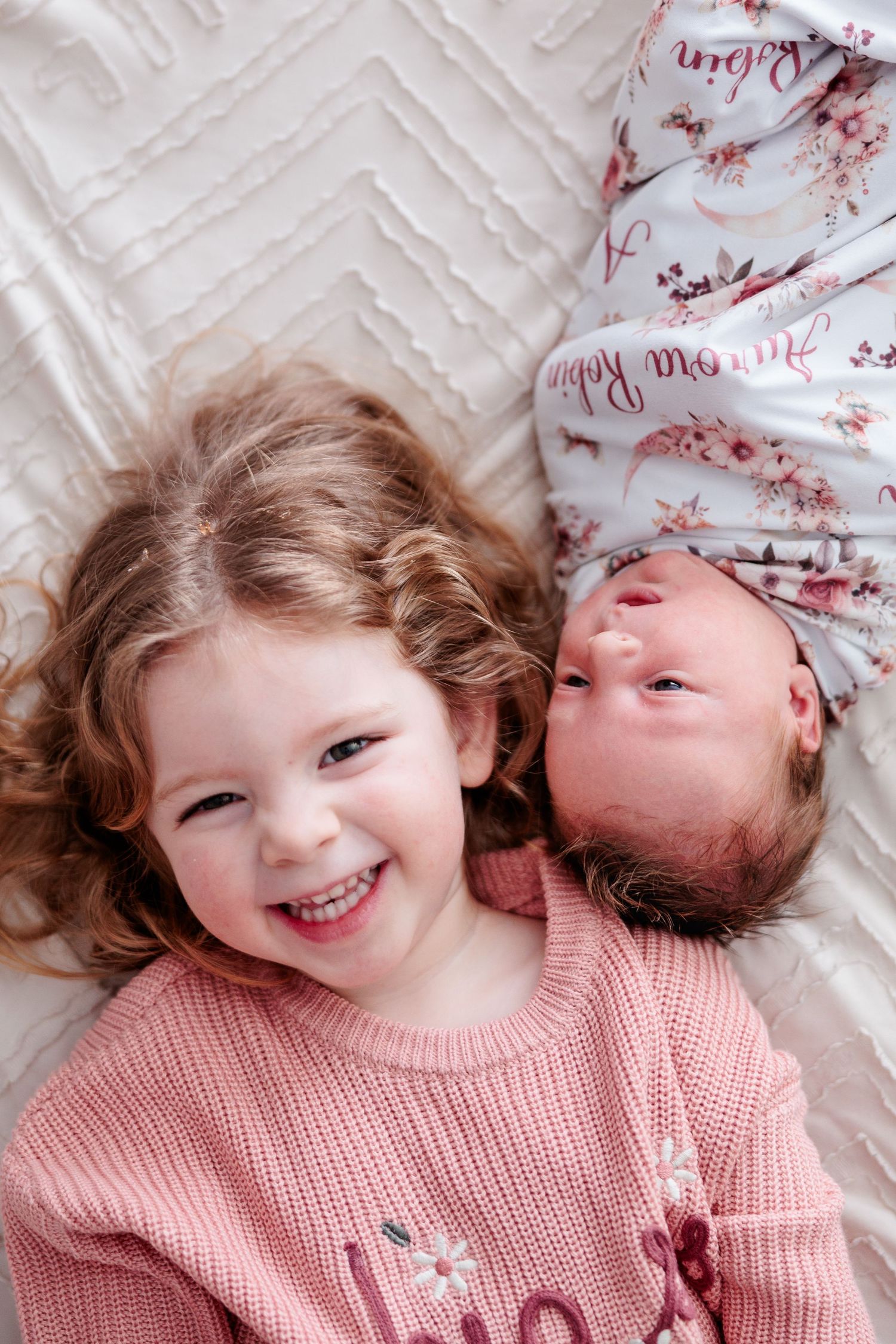 Smiling toddler girl in pink sweater lying next to newborn sibling wrapped in floral blanket.