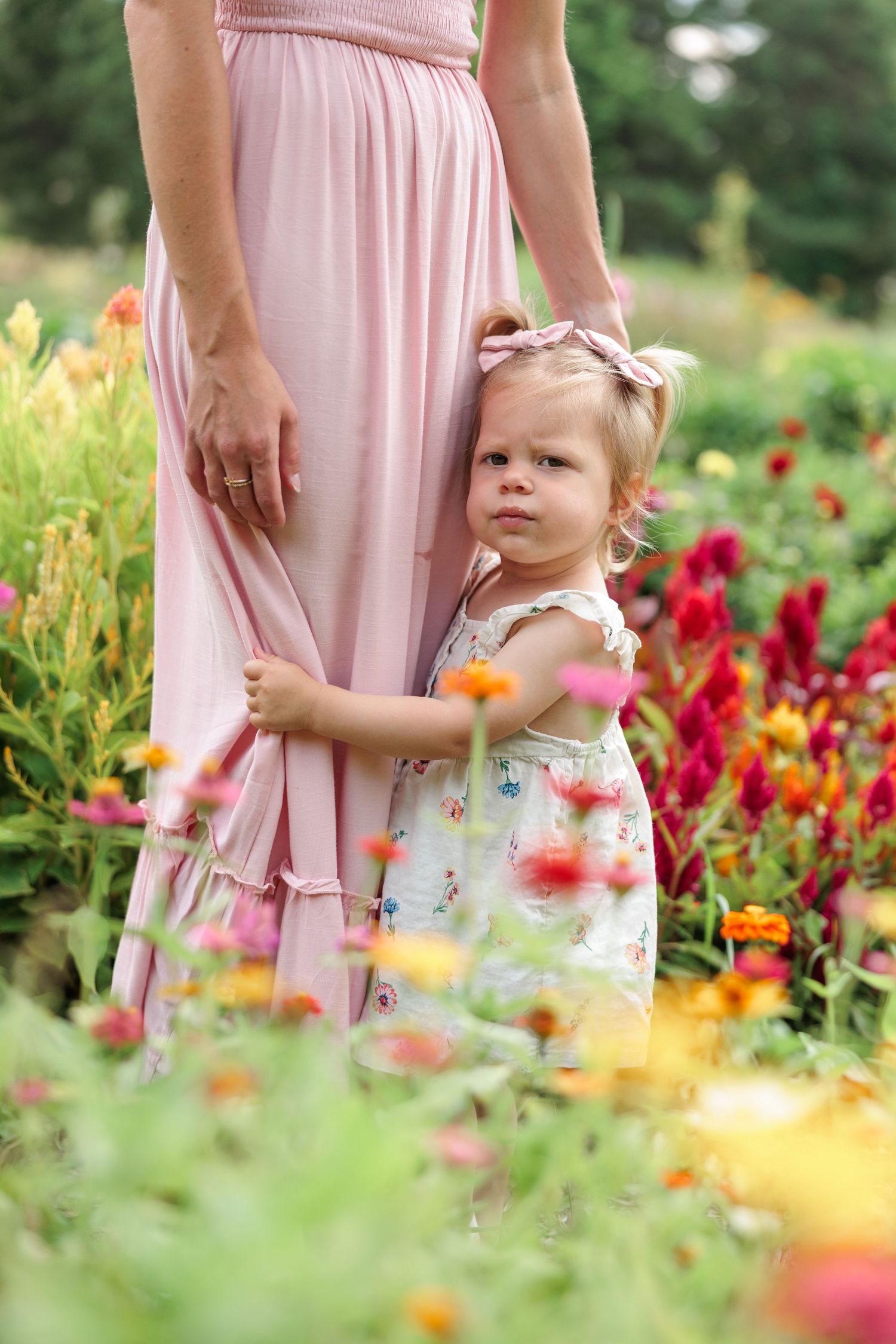A toddler girl in a floral dress clings to her mother's pink skirt in a flower garden.