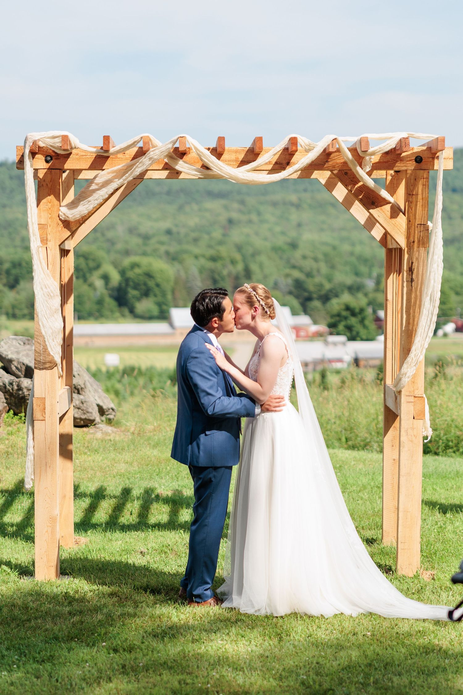 Bride and groom sharing first kiss under a wooden arch with burlap draping at outdoor barn wedding.