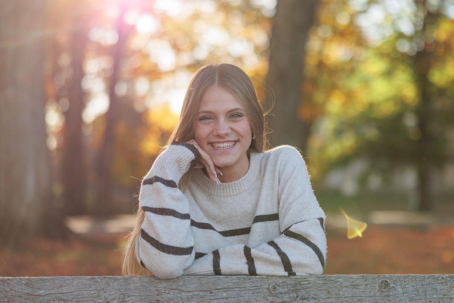 Smiling teenage girl in striped sweater leaning on wooden fence in autumn park with golden sunlight.