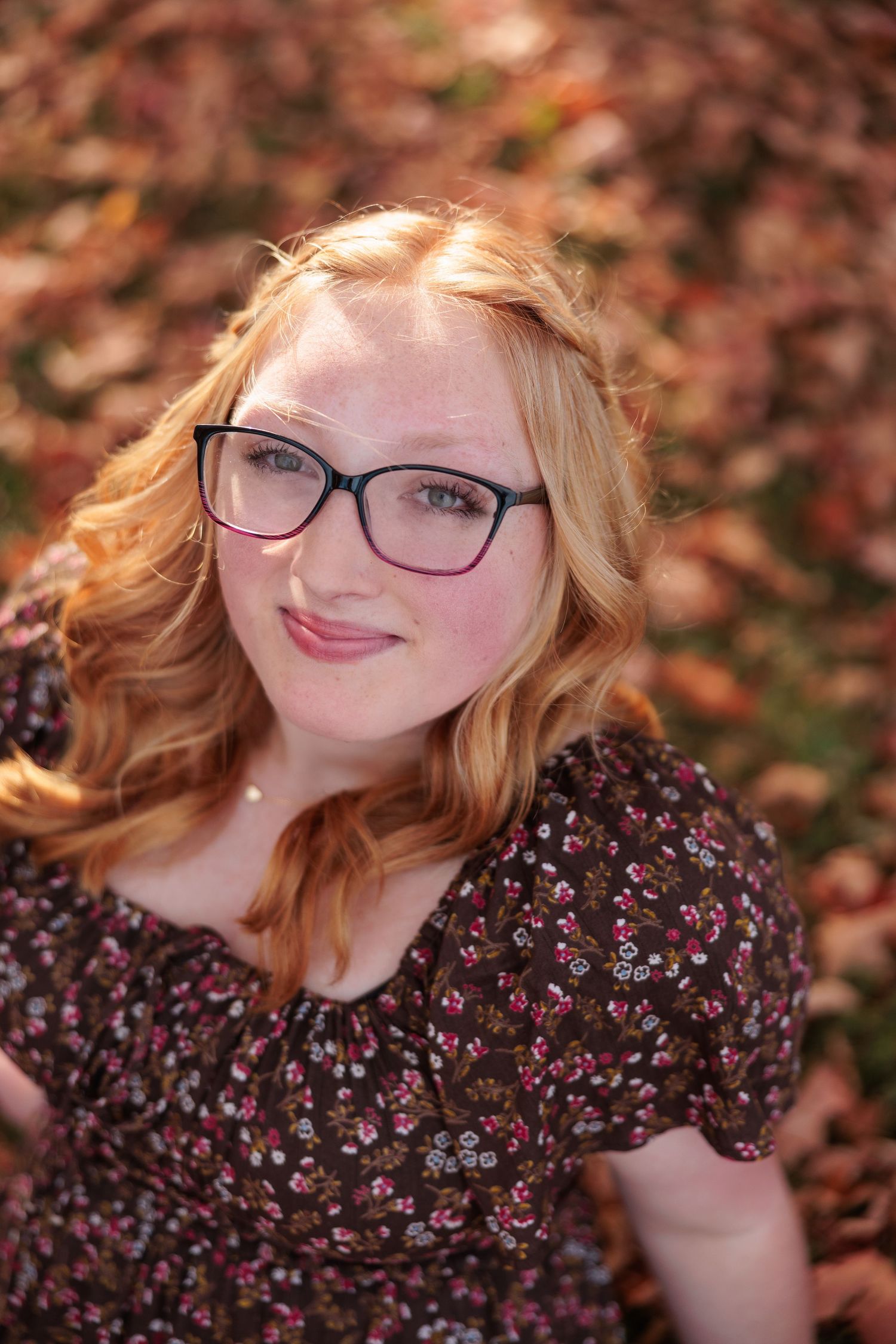 Young woman with red hair and black glasses smiling outdoors among autumn leaves in a floral dress.