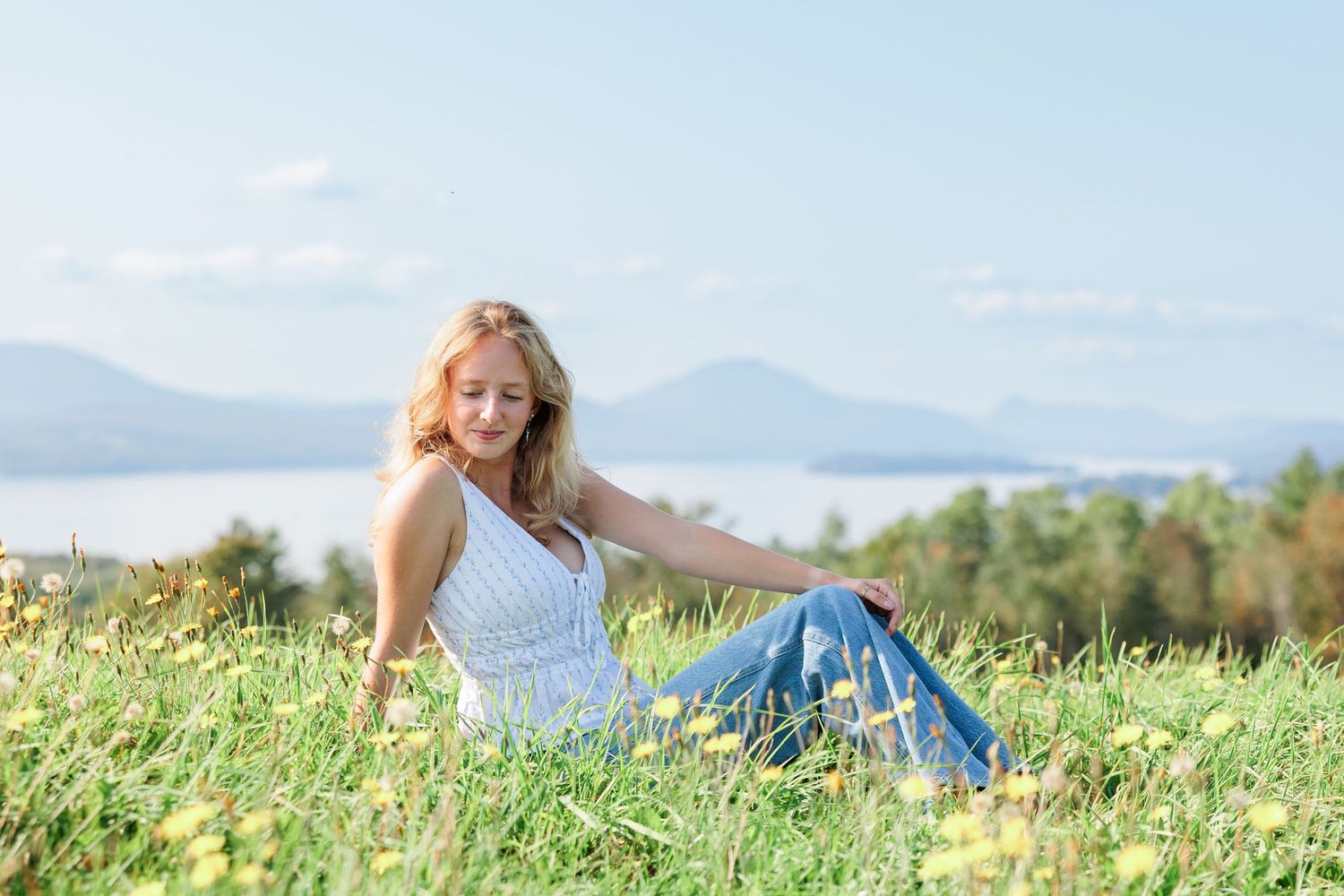 Woman in white top and blue skirt sitting in a sunny green meadow with yellow flowers and mountain lake views.