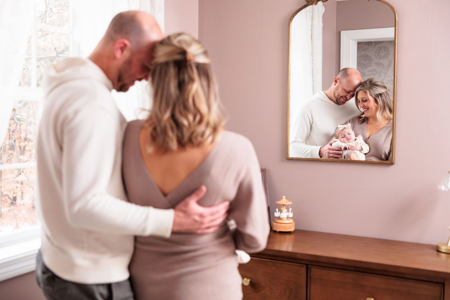 Couple embracing near window reflected in mirror holding newborn baby in softly lit pink nursery.
