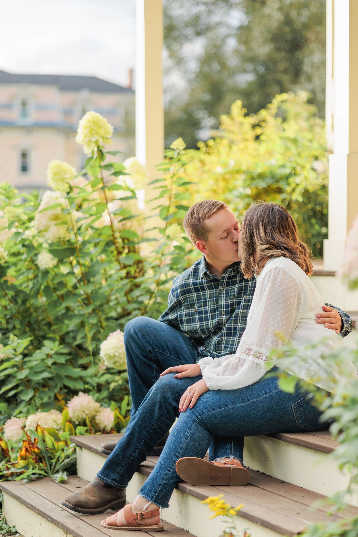 Couple sharing a romantic kiss on porch steps surrounded by blooming hydrangeas and greenery.