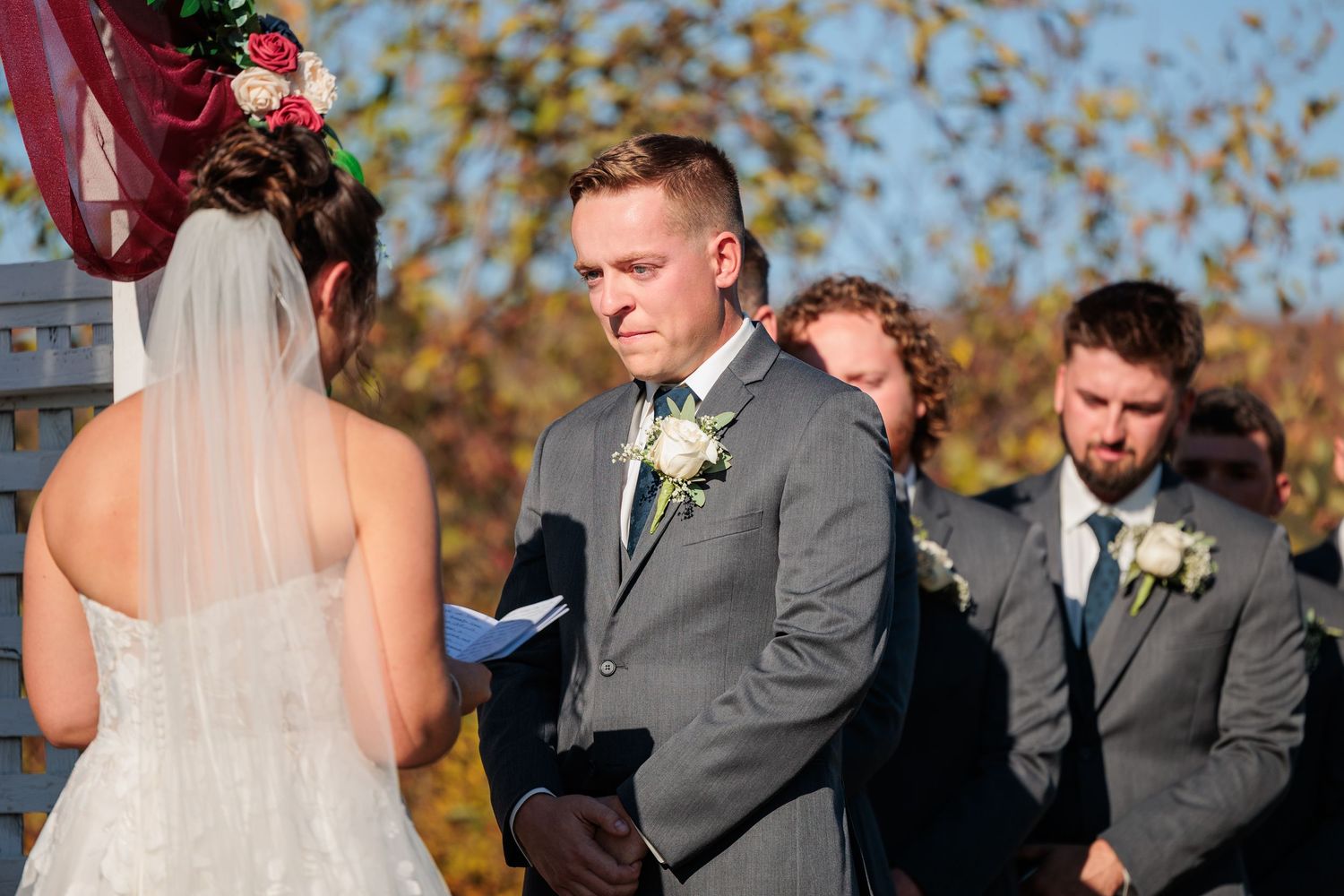 Emotional groom in gray suit listens to bride read vows at outdoor fall wedding ceremony.