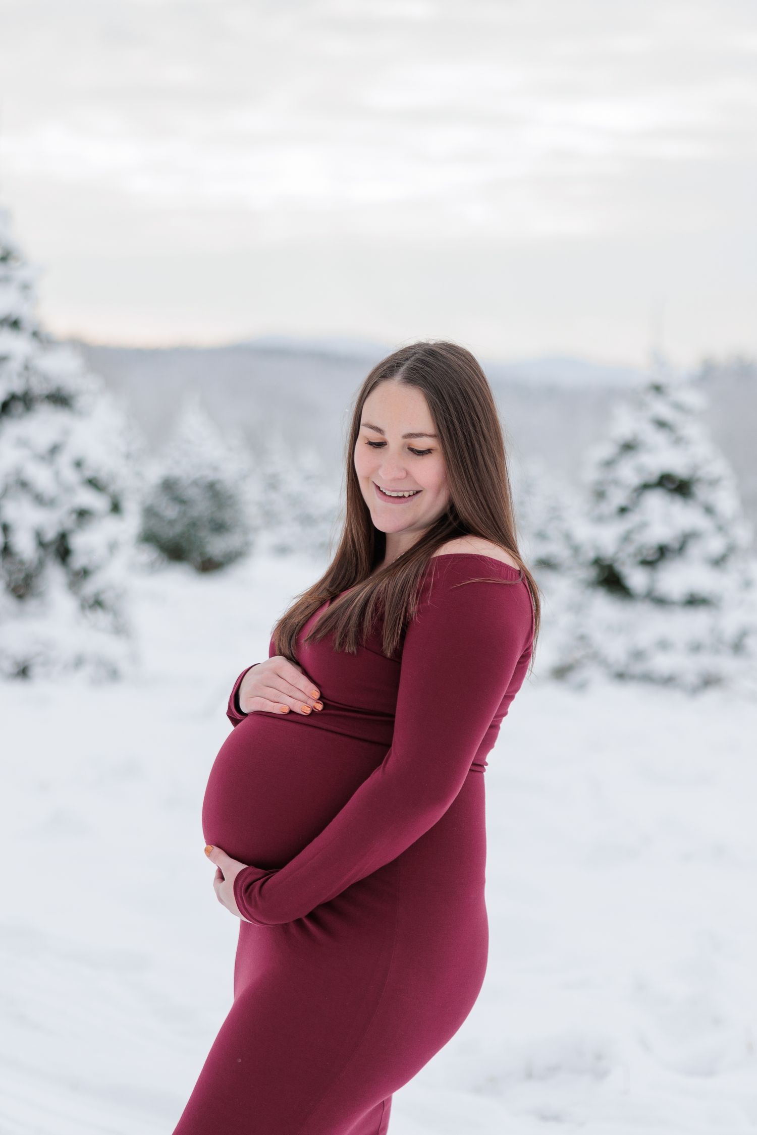 A glowing pregnant woman in a burgundy dress smiles down at her baby bump in a snowy setting.