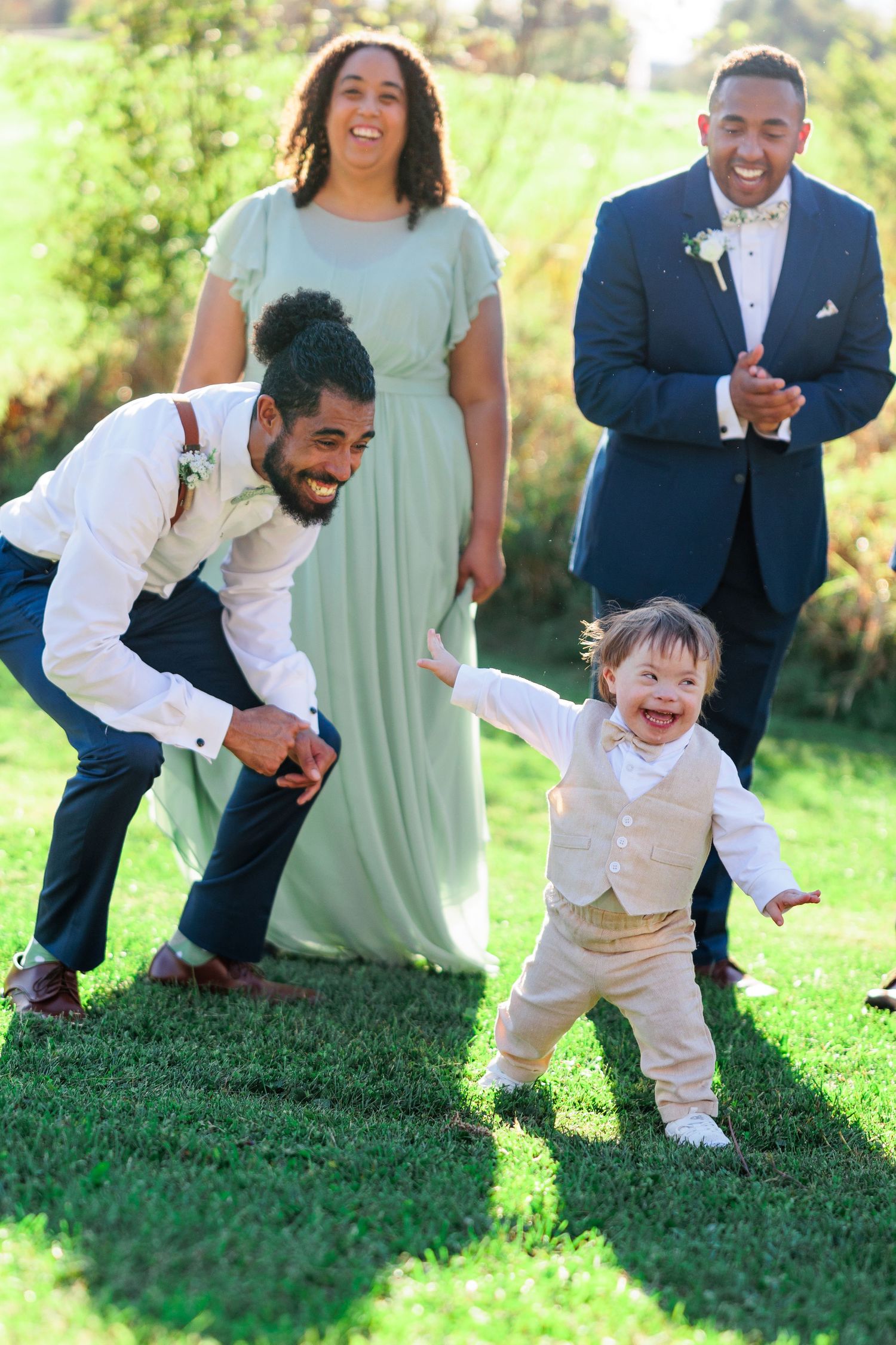 A joyful toddler in a light colored suit plays on green grass during a wedding.