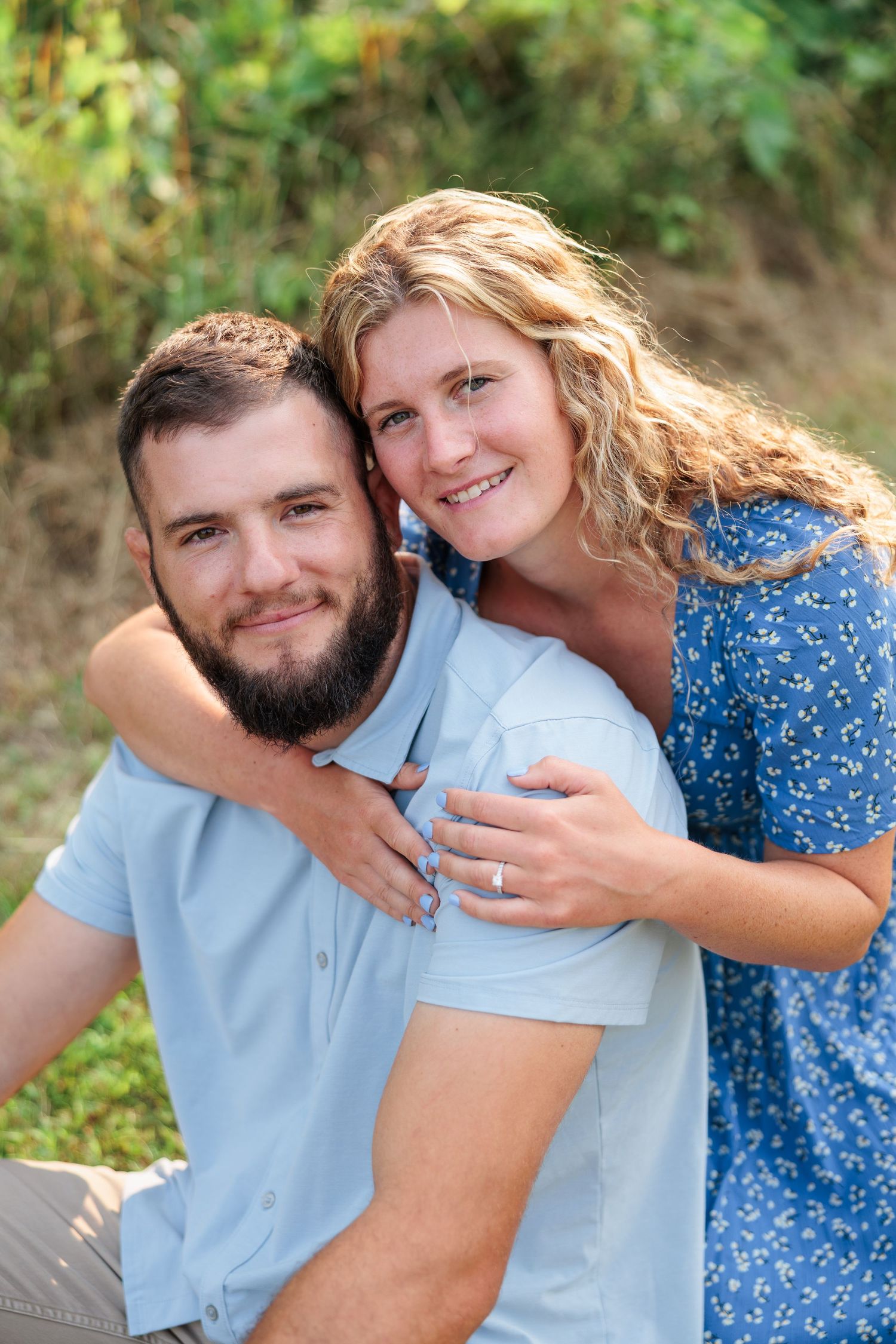 Smiling couple posing outdoors, woman in blue dress hugging man from behind near green foliage.