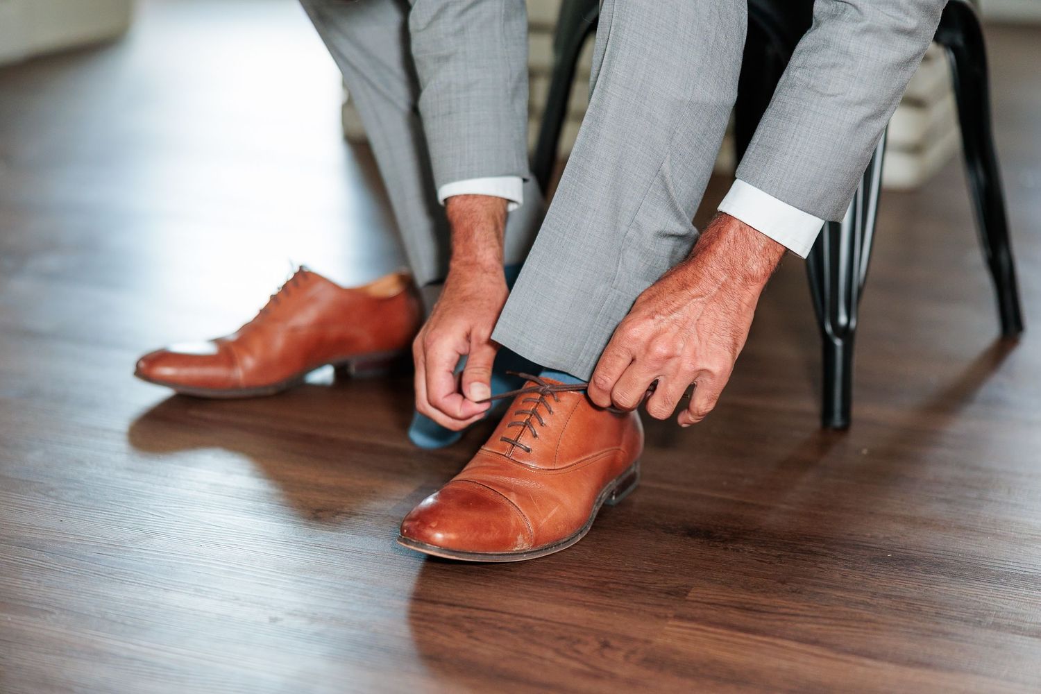 Groom tying brown leather oxford dress shoes while seated on black chair before wedding ceremony.