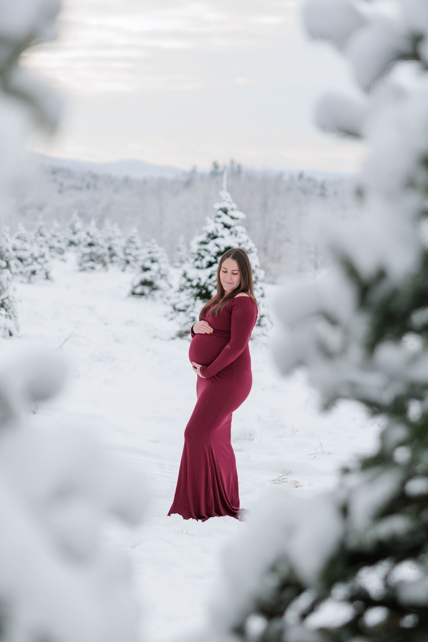 A pregnant woman in a burgundy gown cradles her baby bump among snow-covered evergreen trees.