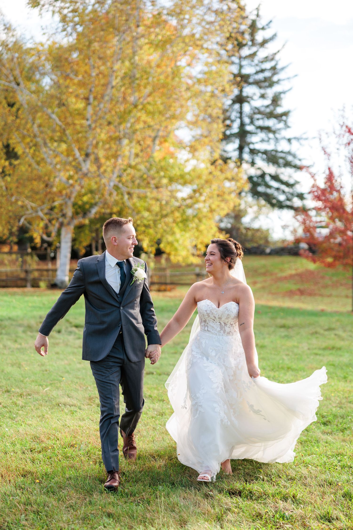 A newlywed couple walks hand in hand through an autumn field with golden trees in the background.