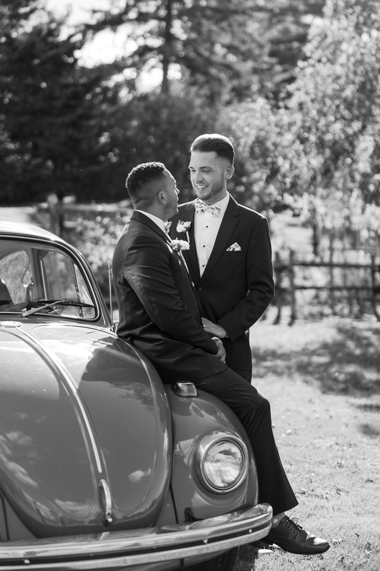 Couple in formal wear pose playfully near vintage car in black and white photography.