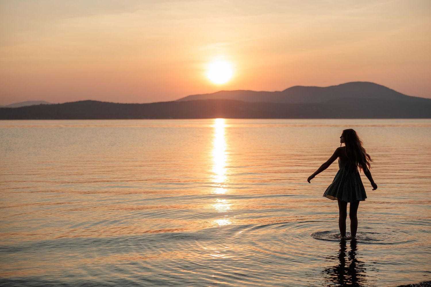 Silhouette of a woman standing in calm ocean water during a vibrant sunset.