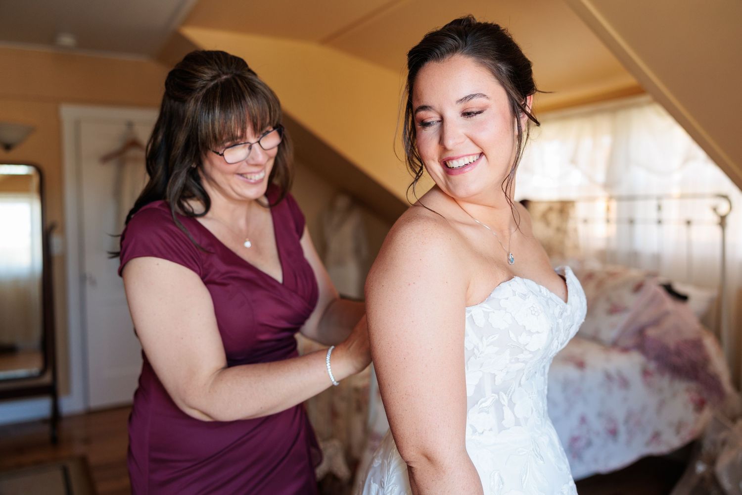 Woman in purple dress helping smiling bride fasten her white lace wedding gown.
