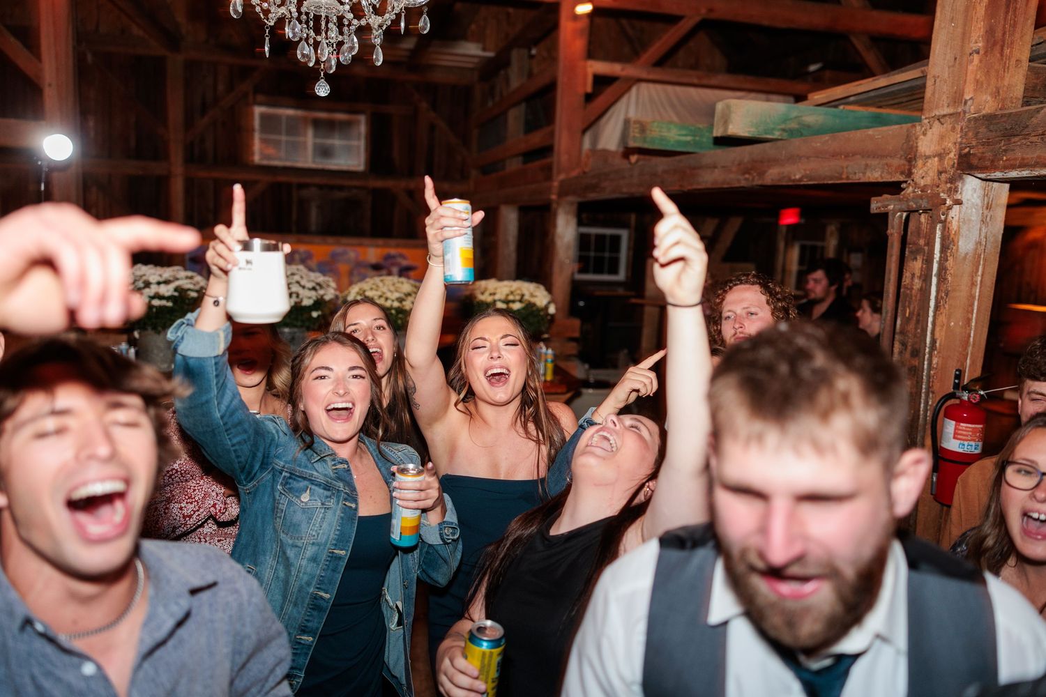 Joyful wedding guests cheer and raise drinks on the dance floor of a rustic barn reception.