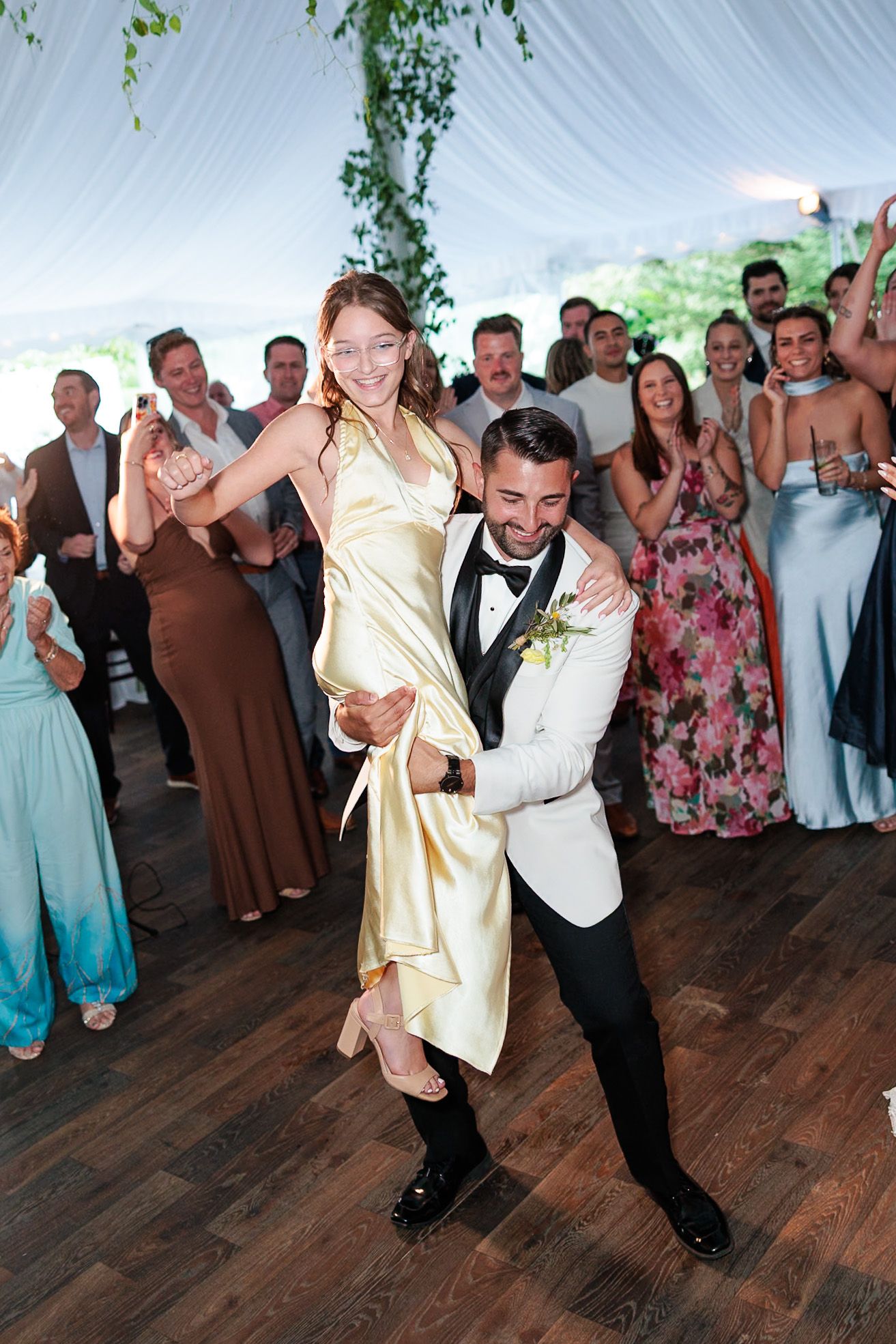 Groom in white tuxedo lifts smiling woman in gold dress on dance floor surrounded by cheering wedding guests.