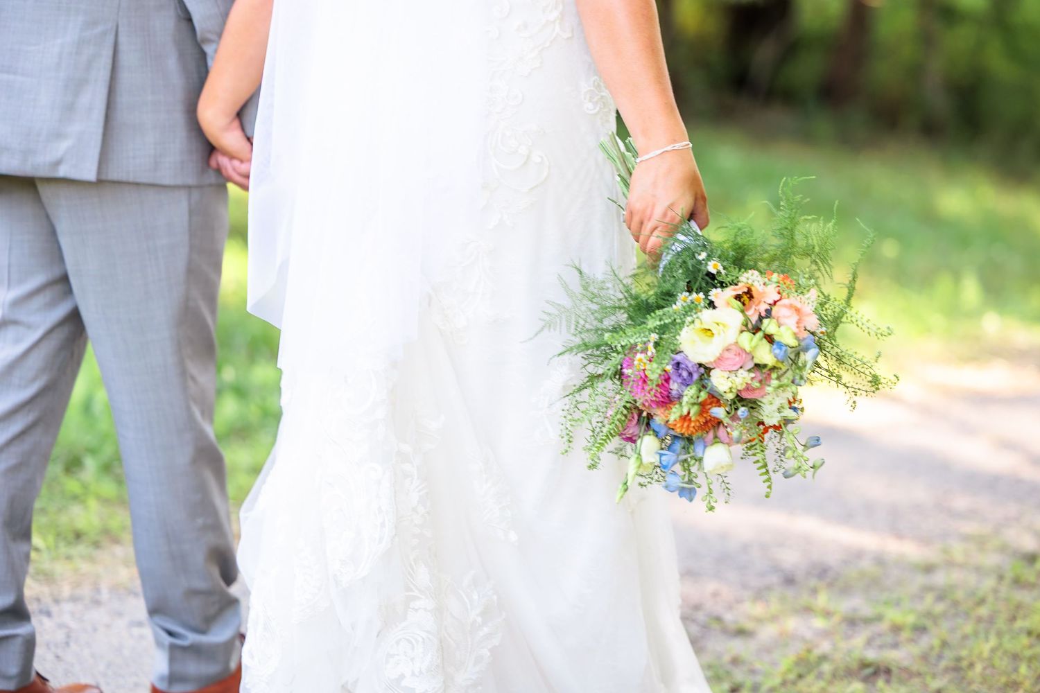 Close-up of bride holding a vibrant wildflower bouquet while standing beside groom in gray suit outdoors.