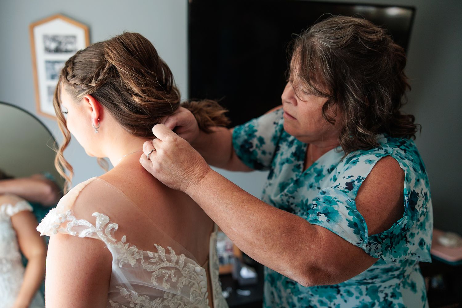 Woman helping bride with lace wedding dress buttons and braided updo hairstyle before wedding ceremony.