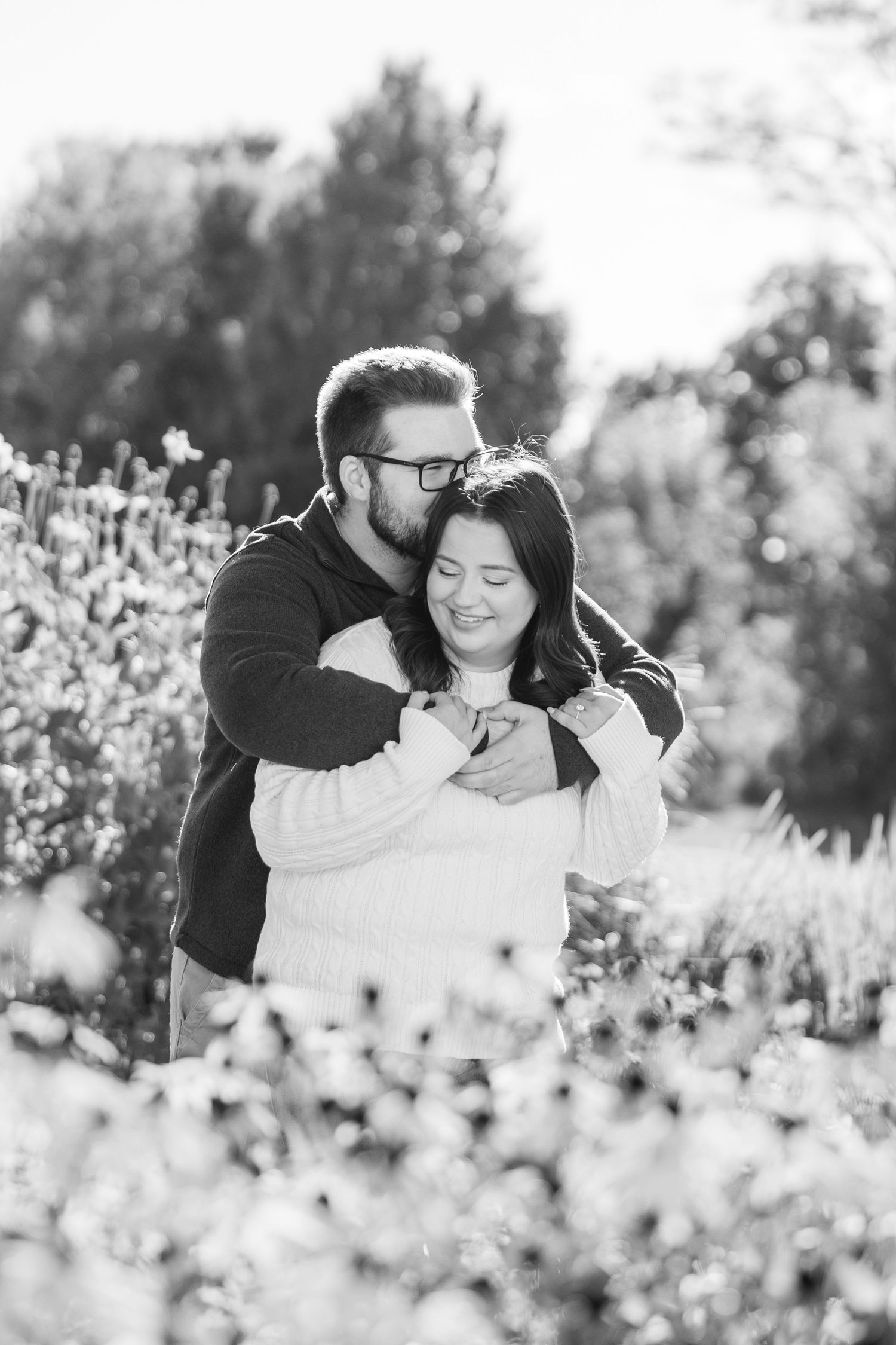 Black and white photo of a man embracing a smiling woman from behind in a garden setting.