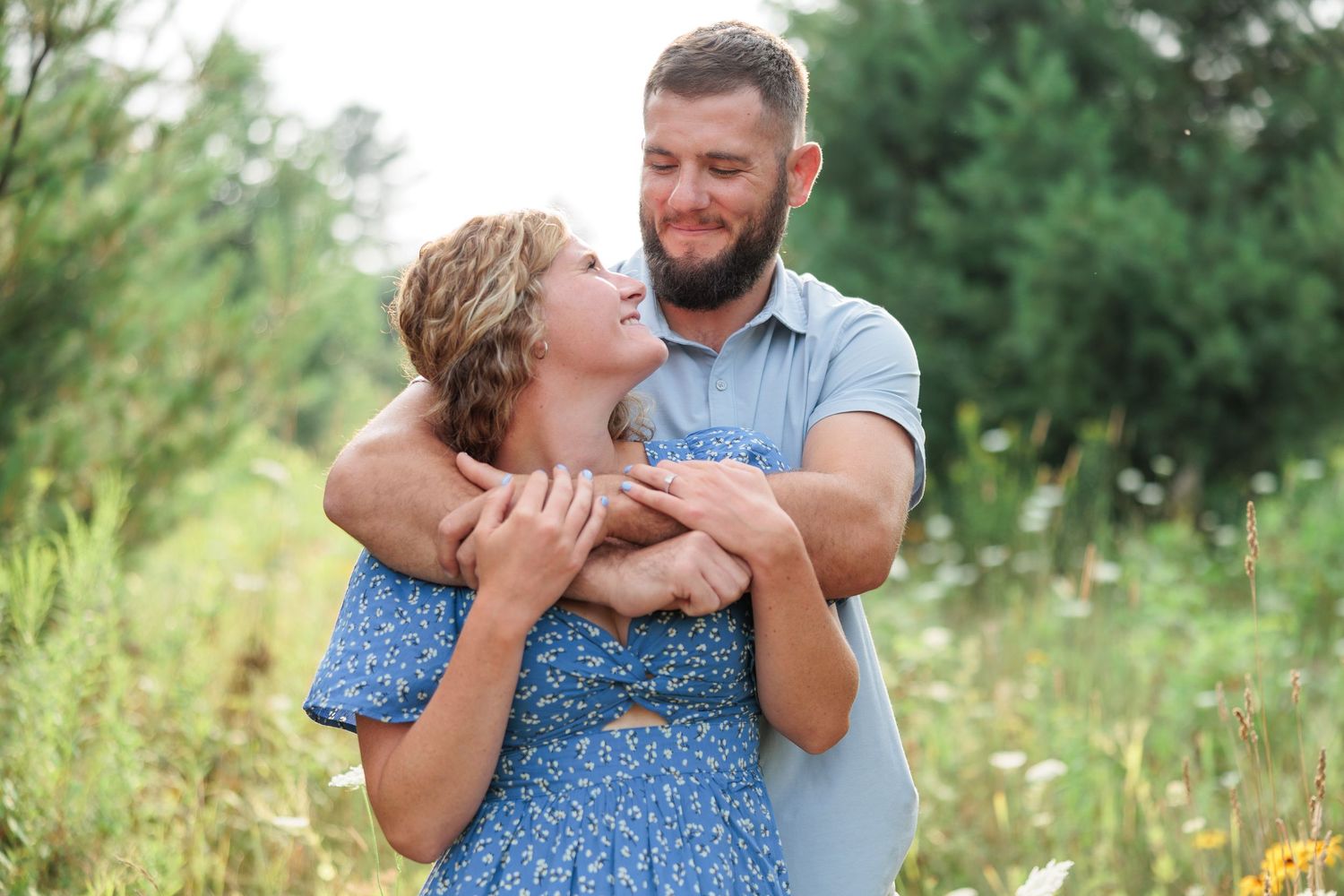 Couple embracing in a wildflower meadow, man hugging woman from behind while she laughs joyfully outdoors.