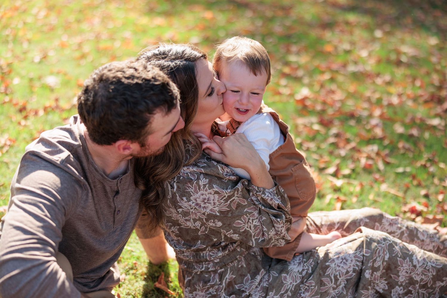 Happy family of three with parents kissing baby boy on cheeks while sitting on fall leaves.