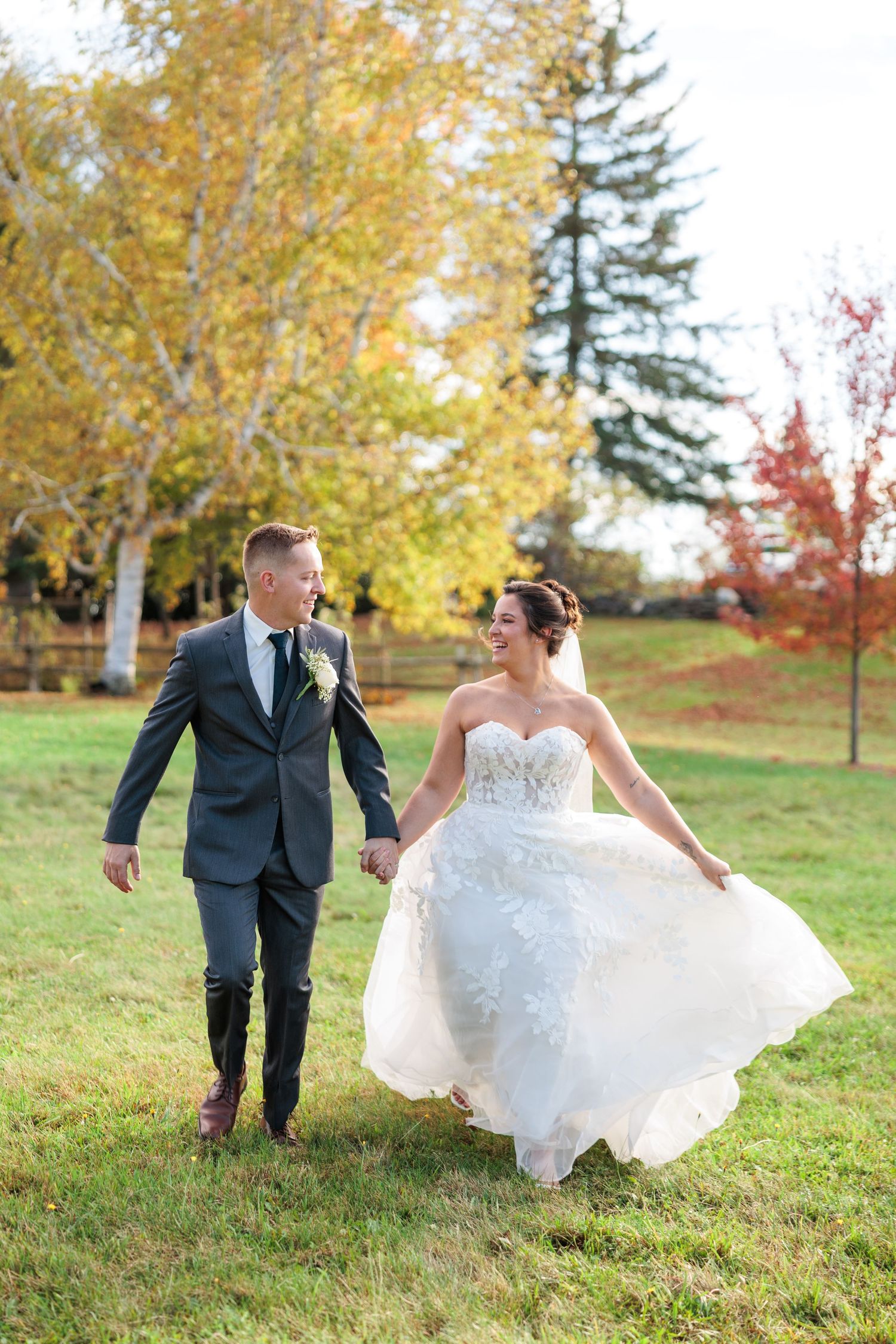 A couple in wedding attire playfully runs and dances together on a grassy hill during autumn with colorful fall foliage.