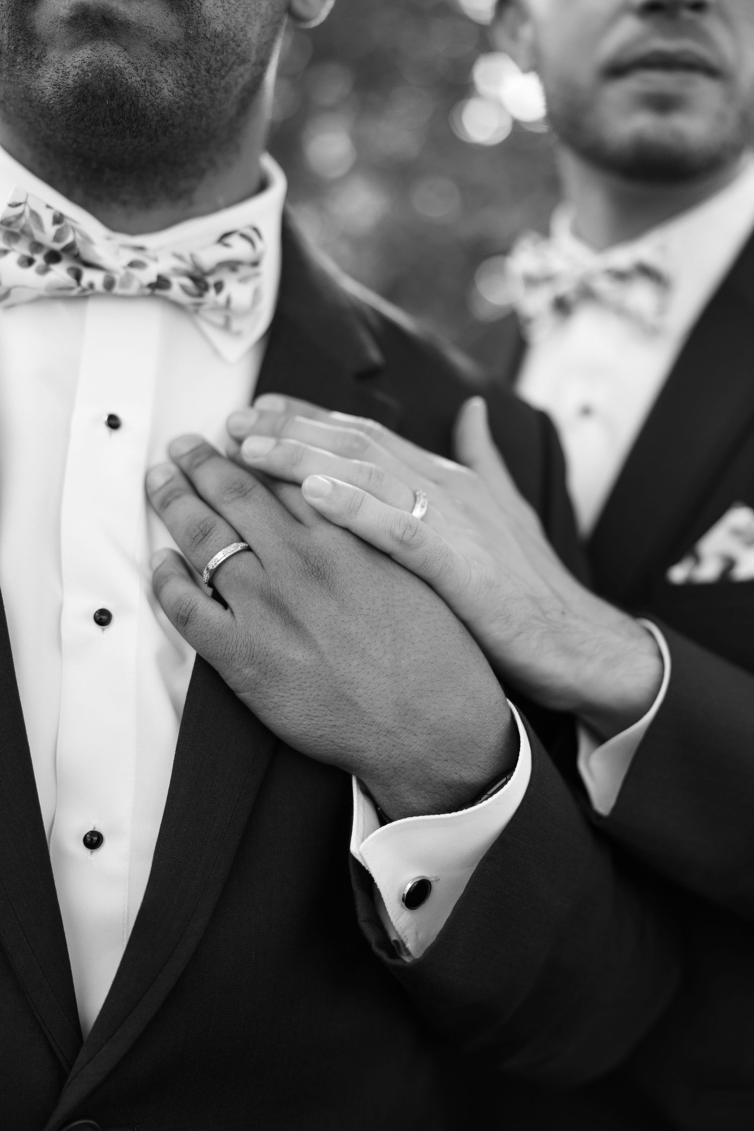 Close-up black and white photo of formal tuxedos with floral bow ties and wedding rings.